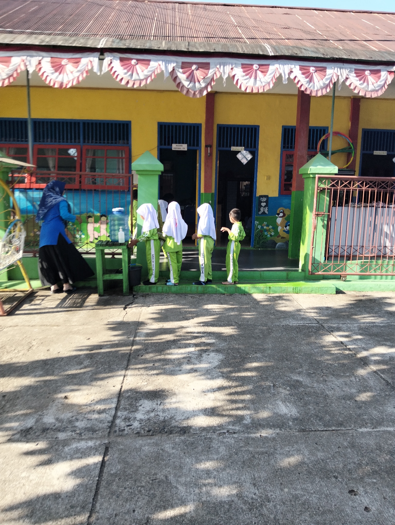 An outdoor, eye-level, full shot captures a group of children in light green uniforms and white hijabs standing in a line on a green, elevated walkway in front of a yellow, two-story building. The building has red-trimmed blue-shuttered windows and a red-tiled roof. A woman in a blue tunic and black skirt, wearing a dark blue hijab, stands to the left, near a green table with a water dispenser and a bottle of hand sanitizer. A young boy in a light green uniform is also present, standing slightly apart from the other children. The ground in front of the walkway is paved with concrete, and dappled shadows from trees are visible on the surface. The scene appears to be in a school setting, likely in Janju, Indonesia, given the style of the building and the attire of the individuals.