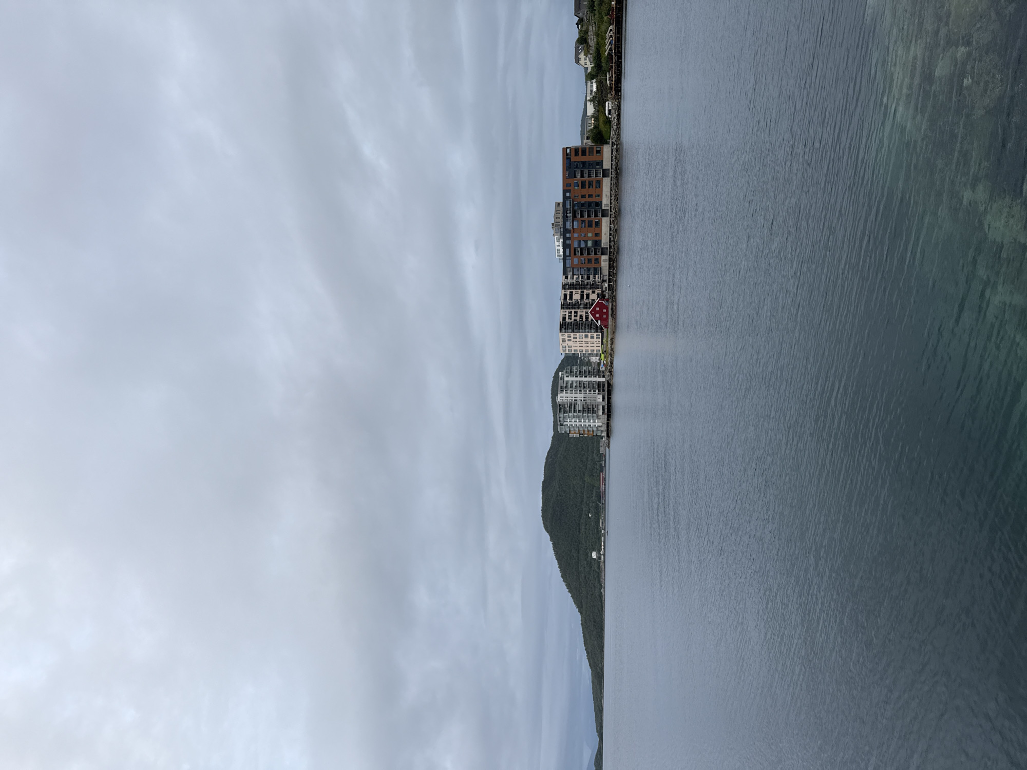 The image shows a waterfront view in Harstad, Norway. In the foreground, a calm, dark blue body of water fills the lower half of the frame. The water appears clear in some areas near the bottom, revealing the seabed. Across the water, in the middle ground, lies a shoreline with a mix of modern and traditional buildings. Dominating the right side of the shoreline are several multi-story apartment buildings with a contemporary architectural style, featuring balconies and various colored facades. To the left of these buildings, a small, bright red, traditional Norwegian house stands out. Further back, a forested hill rises, with some scattered buildings visible at its base. The sky above is overcast with a thick layer of grey clouds, suggesting a cool, possibly damp, day. The overall scene depicts a residential and waterfront area.