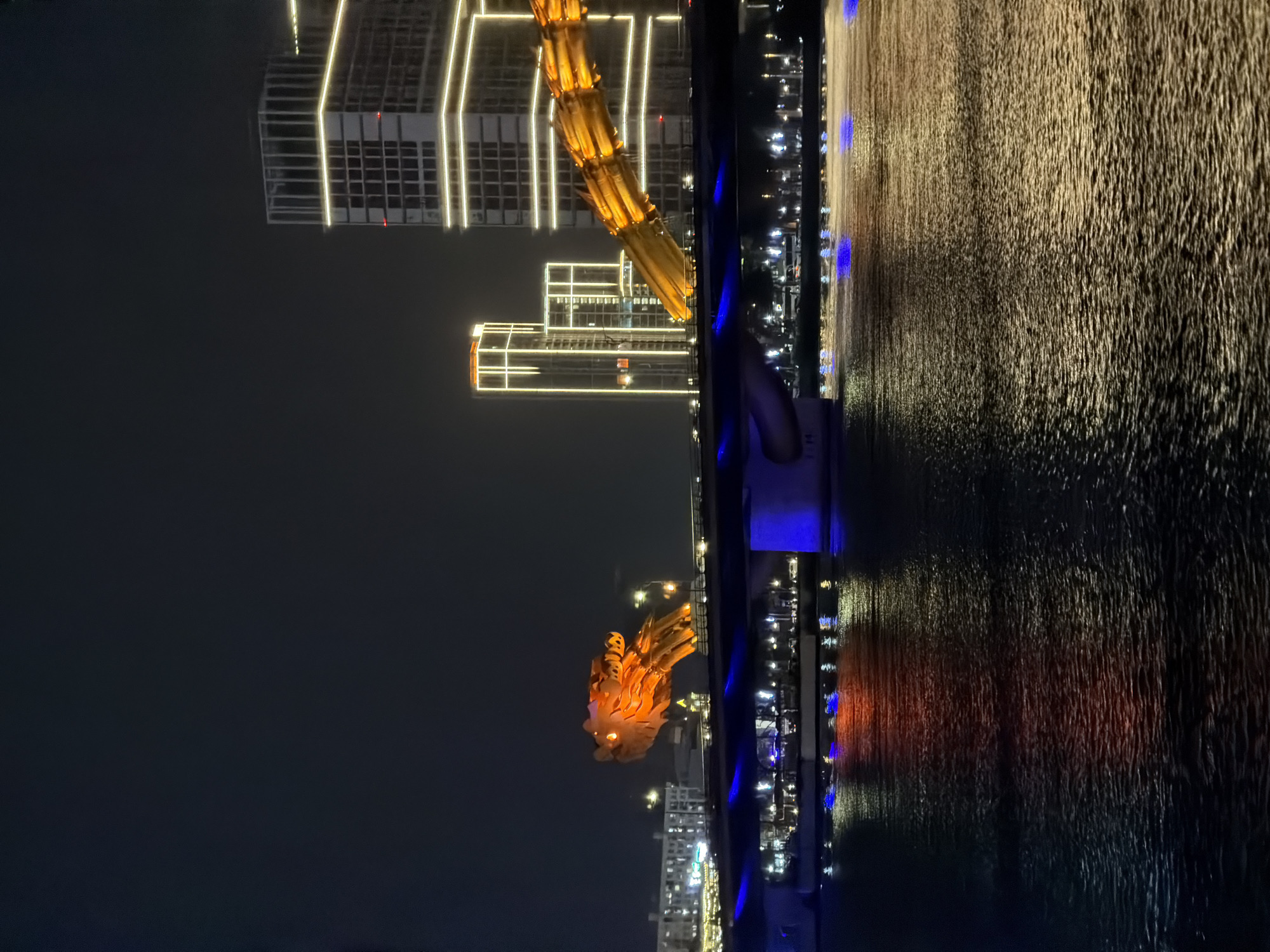 The image displays a vibrant nighttime view of the Dragon Bridge (Cầu Rồng) in Da Nang, Vietnam, spanning across the Han River. The bridge's prominent dragon structure, particularly its head and a substantial part of its body, is brilliantly lit with a golden-orange light, creating a striking focal point. The dragon's eyes appear to glow brightly.
Below the illuminated bridge, the surface of the Han River is alive with reflections. The artificial lights from the city and the bridge itself cast shimmering patterns on the water, appearing as elongated streaks and patches of light, primarily in golden, orange, and blue tones. The bridge structure itself is adorned with distinct blue lights, which run along its length, creating a cool contrast to the warm glow of the dragon.
In the background, several modern high-rise buildings are visible, their architectural lines emphasized by bright, linear white lights that trace their outlines against the dark night sky. The overall scene conveys the bustling and luminous atmosphere of Da Nang at night. There are no people or specific activities clearly discernible, and no legible text is visible in the frame.