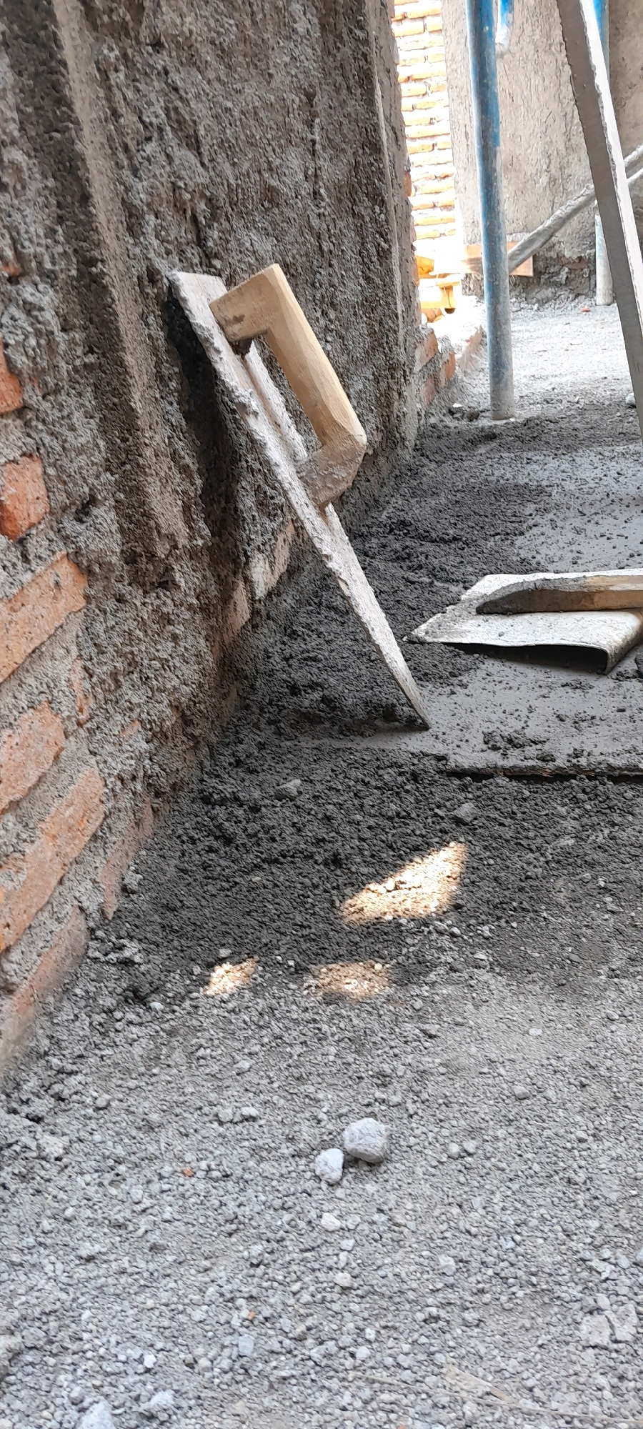 A construction scene is captured in Karangduren, Indonesia. A trowel with a wooden handle is propped against a brick wall covered in wet cement. The ground is composed of gravel and wet cement. In the background, scaffolding and more brickwork are visible. The lighting suggests it is daytime, with shadows indicating sunlight.