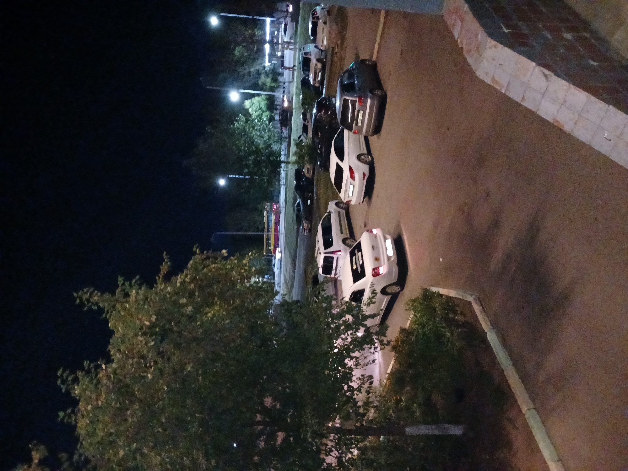 This is a nighttime aerial shot of a parking lot in Kostanay, Kazakhstan, likely near a residential or commercial building given the presence of a tree in the foreground and a brick structure on the right. The parking lot is occupied by several cars, including a white sedan in the foreground, a silver hatchback behind it, and various other vehicles further back. Streetlights illuminate the scene, casting long shadows. In the background, a yellow and red bus is visible on a road, suggesting the proximity of a transportation route. The environment appears urban, with paved surfaces and some greenery.