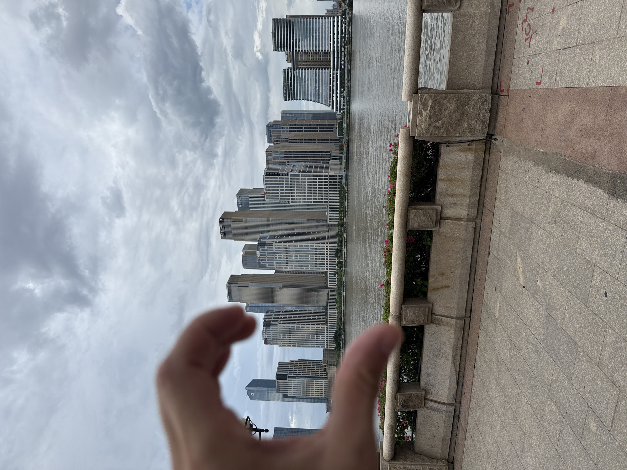 A hand forms a heart shape in the foreground, obscuring a portion of the view of a waterfront cityscape. In the background, a river flows, and across the river, a line of modern skyscrapers with glass facades and varied architectural designs dominate the skyline. The sky is overcast with clouds. The foreground features a paved walkway and a stone balustrade with some greenery and pink flowers planted behind it. The scene appears to be a public space, likely a promenade or park area along a river. The location is identified as Tianhe District, China.