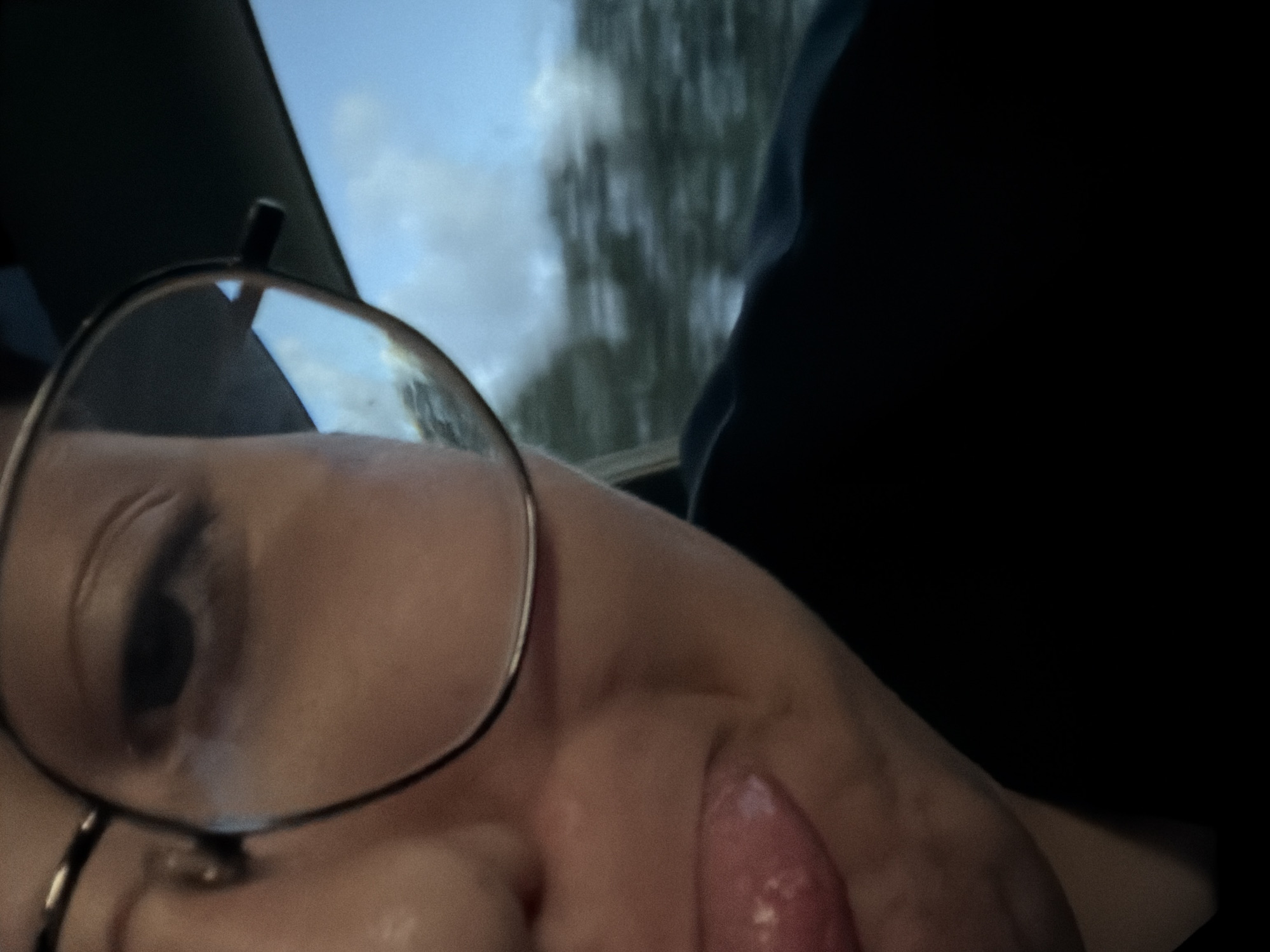 The image is a close-up, self-taken photograph of a person wearing glasses. The person's face is partially visible, with one eye and the mouth in focus. The person is sticking their tongue out. The background shows a blurred view of trees and the sky, suggesting the person is likely in a vehicle moving through a landscape. The lighting is overcast, indicating it is likely daytime. The image does not contain any specific visual cues that place it in Moss, Norway.