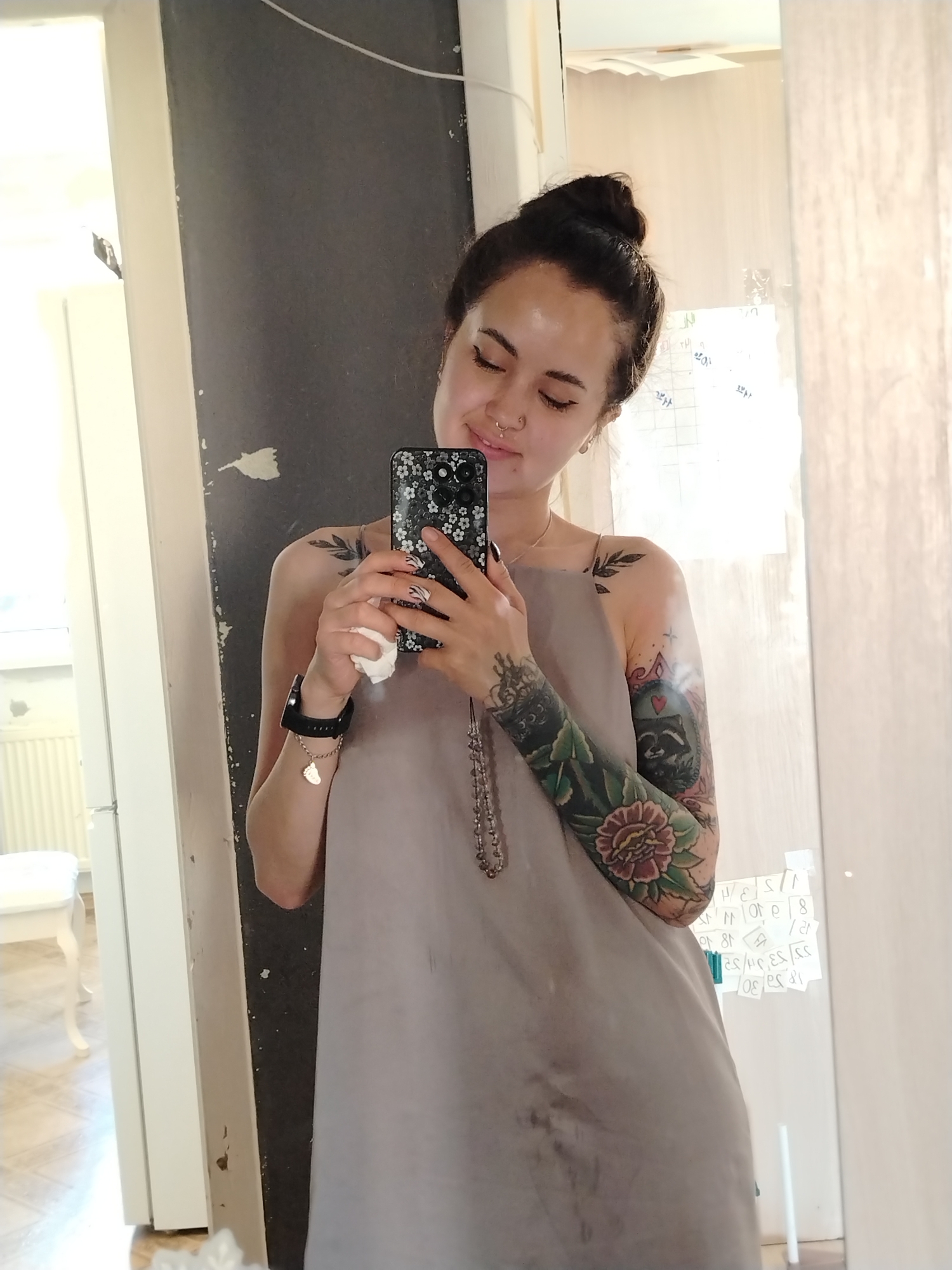 A young woman takes a selfie in what appears to be an indoor setting. She has her hair styled in a bun and has visible tattoos on her arms and shoulders. She is wearing a loose-fitting gray dress. She is holding a smartphone with a floral-patterned case, taking a picture of herself in the mirror. The background shows a partially visible room with light-colored walls and furniture. There is a window visible behind her. A whiteboard with notes is also present. The location in the city of Noviy, Russia is not indicated in the image.