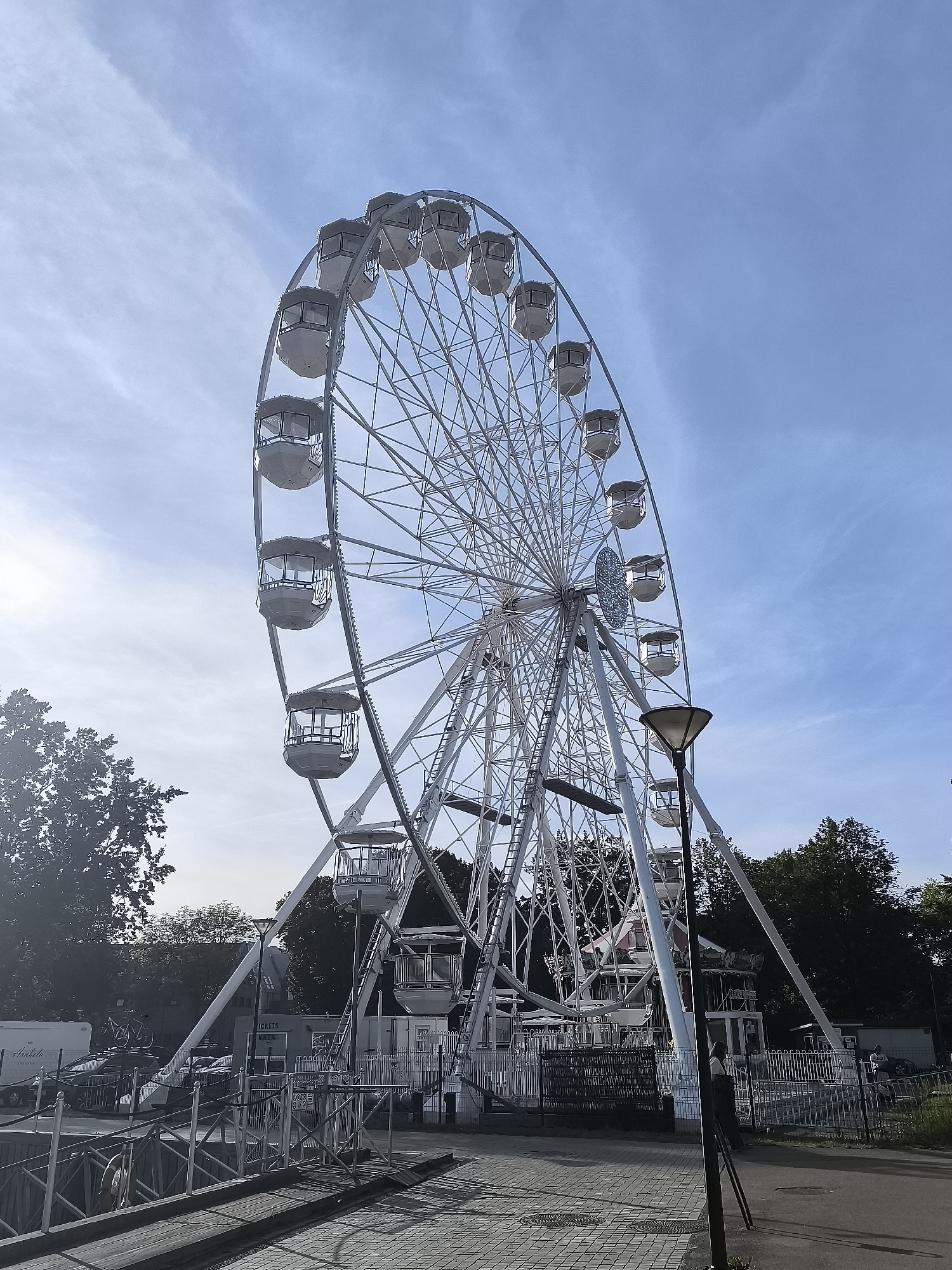 The image shows a large white Ferris wheel against a bright blue sky with scattered clouds. The Ferris wheel has multiple white gondolas attached to its circumference. In the foreground, there is a paved area with patterned tiles, a metal fence, and some trees. Behind the Ferris wheel, more trees and some buildings are visible. The scene appears to be an amusement park or fairground. The context of Klaipėda, Lithuania, suggests this is a scene from that city.