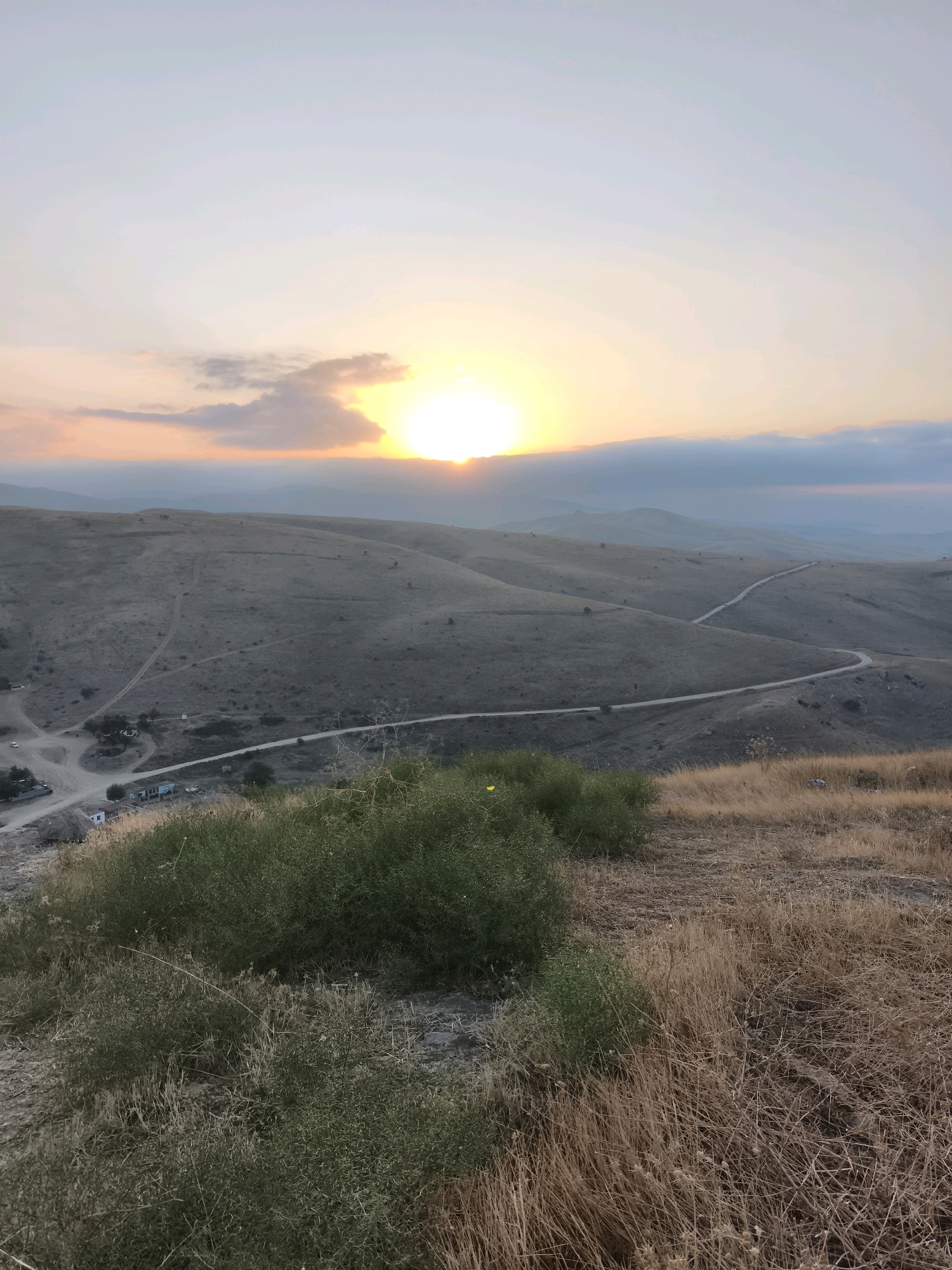 The image shows a panoramic view of a hilly landscape at sunset. The sun is setting behind a bank of clouds, casting a warm glow over the scene. The hills are covered in dry grass and scattered bushes. A winding road snakes through the hills, leading to a small settlement in the distance. The settlement appears to consist of a few buildings and some parked vehicles. The sky is a pale blue, with streaks of orange and pink from the setting sun. The foreground of the image is dominated by dry grass and a few green bushes. The overall impression is one of a remote and peaceful rural area. The specific location in Qalaşıxı, Azerbaijan is not discernible from the image alone.