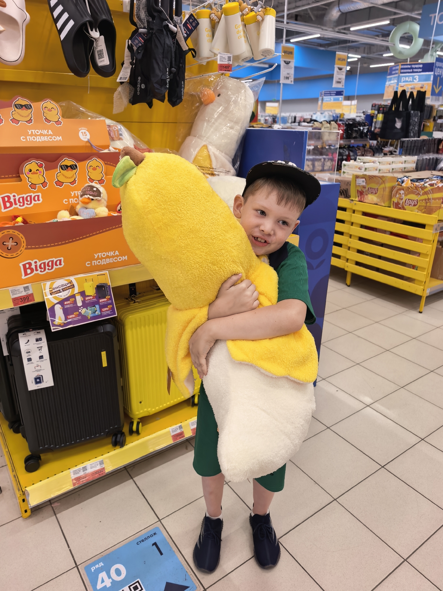 A young boy is standing in a supermarket aisle holding a large plush toy shaped like a banana. He is wearing a green t-shirt and shorts and a black baseball cap. The toy is yellow and white, and its stem is green. In the background, shelves are stocked with various products, including suitcases, chips, and clothing items. A sign on the floor indicates "ряд 40" (row 40) and "стеллаж 1" (shelf 1), suggesting a specific location within the store. The floor is tiled in a light color. The overall environment suggests a retail setting in Sterlitamak, Russia.
