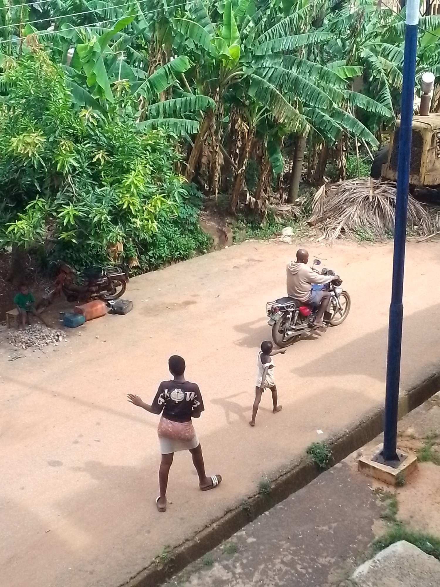 The image depicts a street scene in an unknown location. In the foreground, a person wearing a t-shirt and skirt stands on the edge of the road, with another child walking nearby. In the middle ground, a man rides a motorcycle. In the background, there is lush green vegetation, including banana trees. To the right of the background is a parked, yellow bulldozer. Two boys are also visible on the left side of the image, sitting on the side of the road. There is a blue pole in the right foreground. The scene is bathed in sunlight, creating shadows on the road.