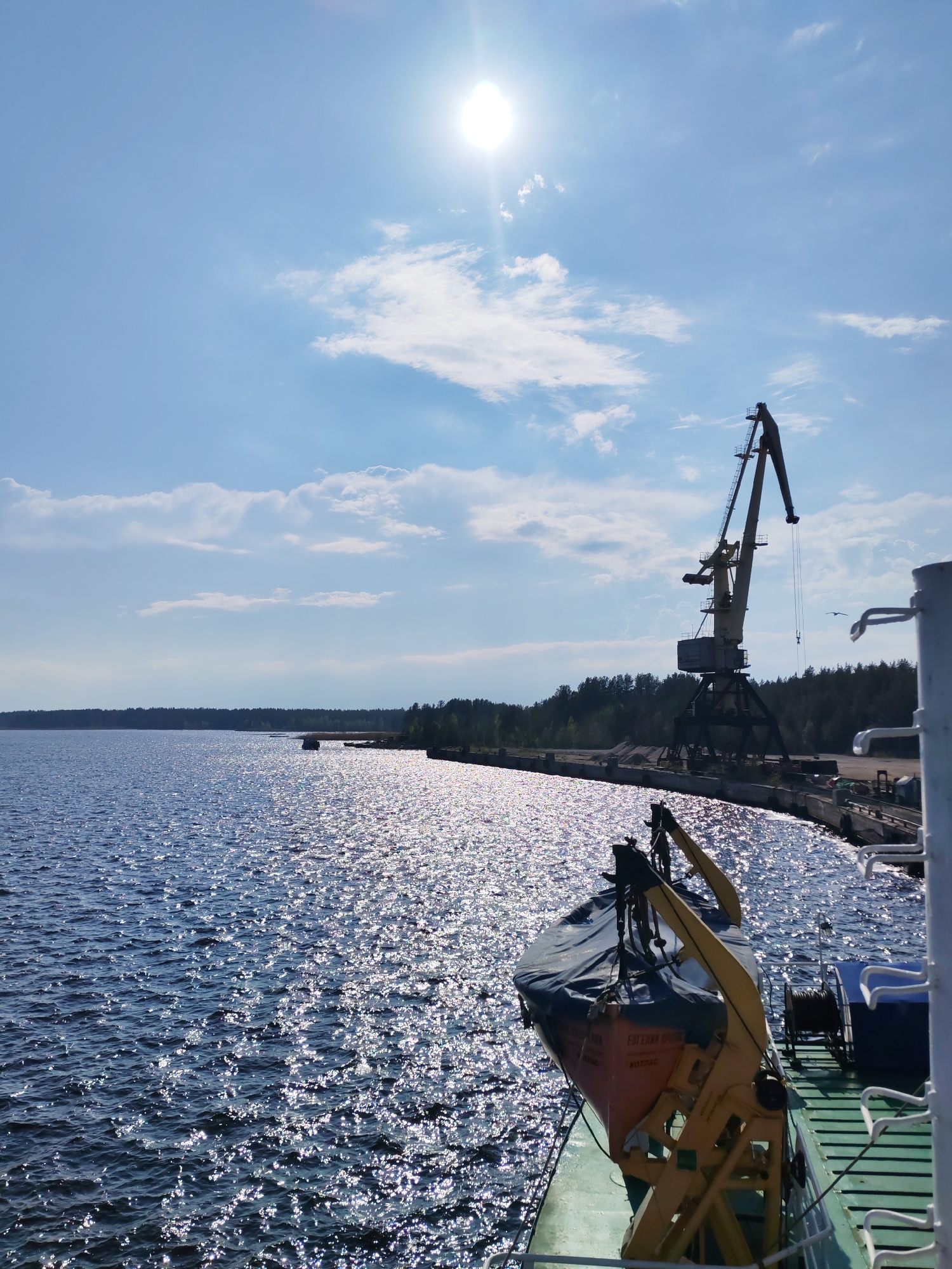The image depicts a bright, sunny day at a waterfront in Shalsky, Russia. A large body of water shimmers with reflected sunlight, filling much of the frame. In the middle ground, a crane stands on a dock, with a line of trees along the coast in the background. In the foreground, part of a ship is visible, including a covered lifeboat in its yellow frame. The sky is blue with scattered white clouds, and the sun shines brightly.