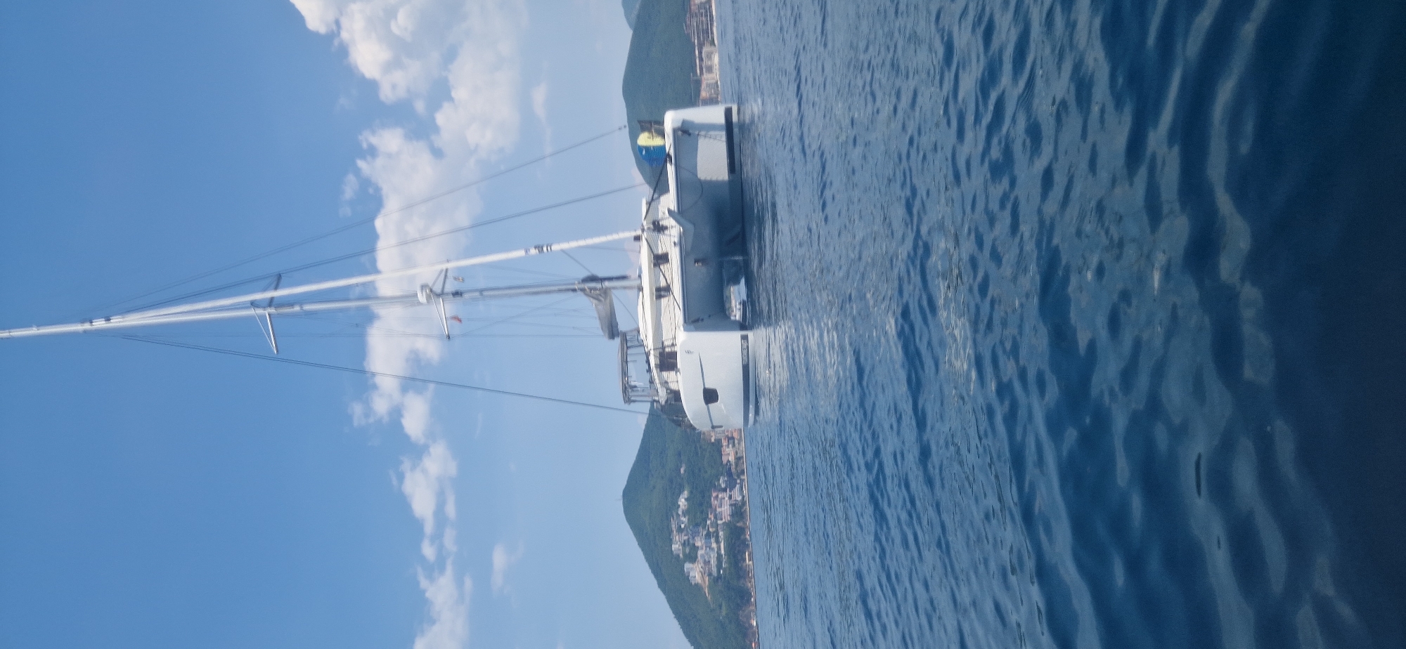 The image shows a white sailboat with a tall mast and rigging on a bright, sunny day. The sailboat is on the water, which appears calm with gentle ripples. In the background, there is a lush green hillside covered with buildings, suggesting a coastal town. The sky is a clear blue with a few fluffy white clouds. The image is oriented vertically, with the mast pointing upwards and the water occupying the right side of the frame. It is difficult to determine the exact time of day, but the bright sunlight suggests it is daytime. There is no visible text in the image.