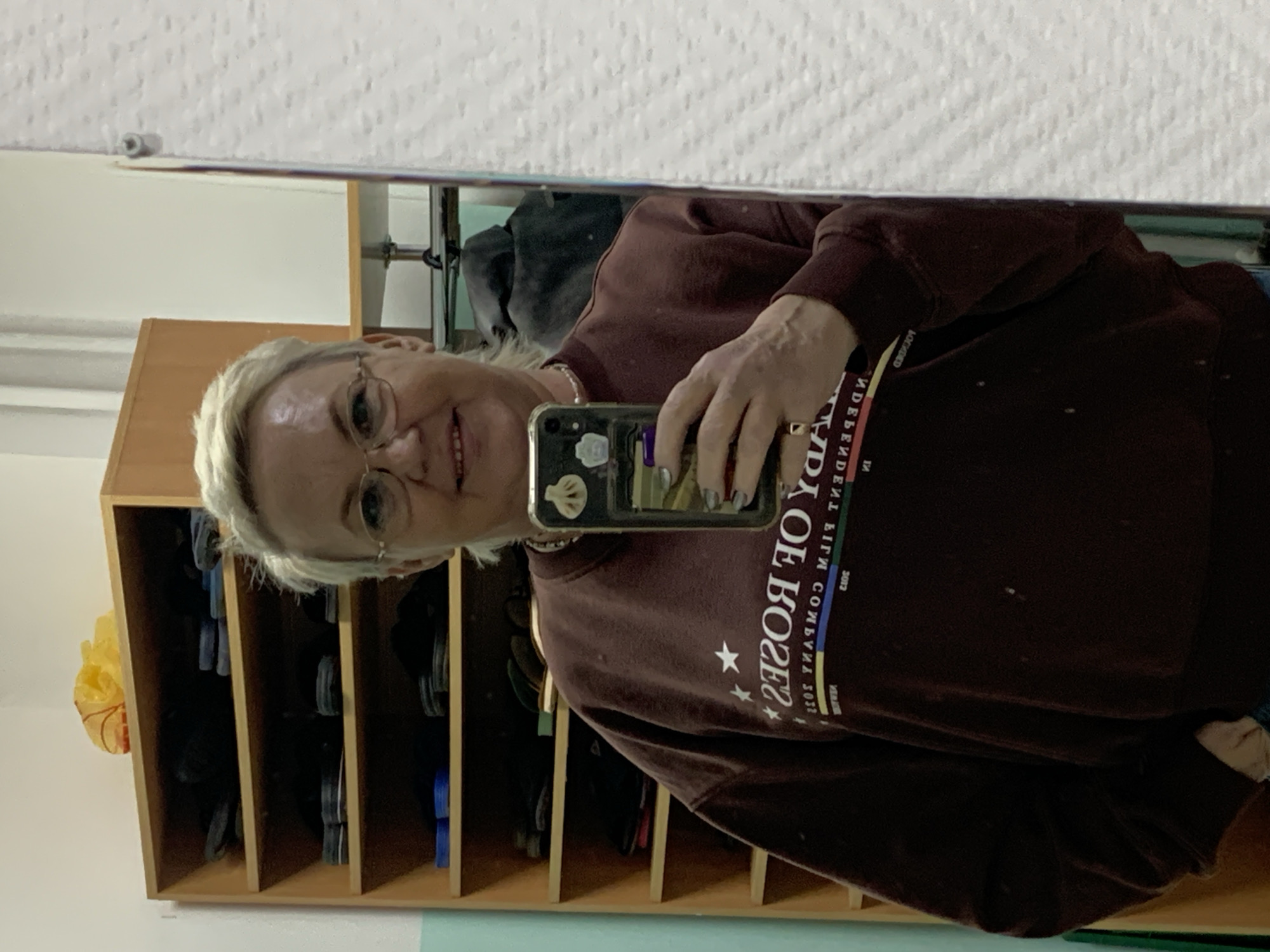 A middle-aged woman with short, blonde hair and glasses is taking a selfie in a mirror. She is wearing a maroon sweatshirt with white lettering. Behind her is a wooden shelving unit filled with various items. The photo appears to have been taken indoors, possibly in Chaikovsky, Russia.