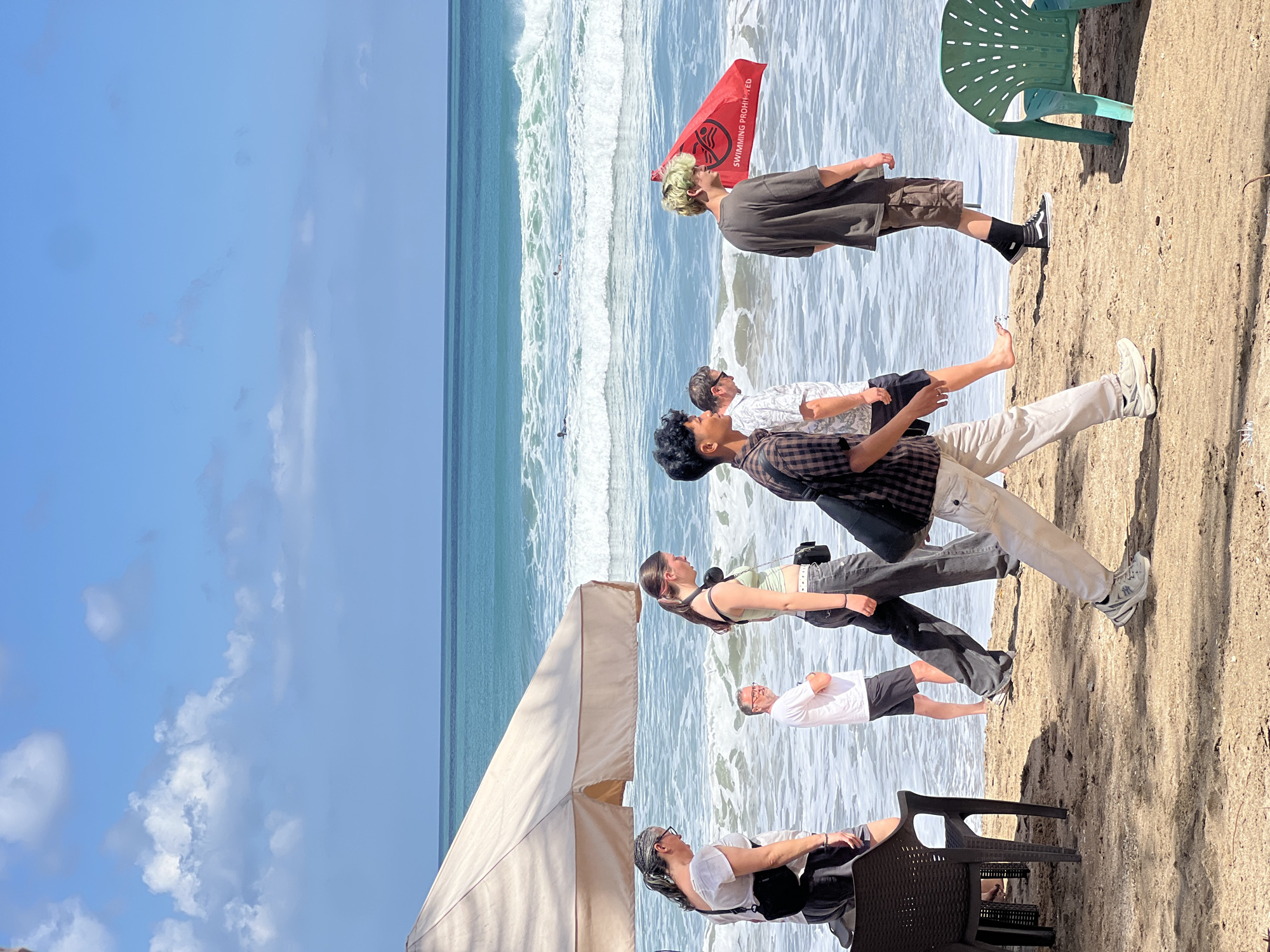 The image depicts a sunny day at Kuta Beach, Indonesia, with several people walking along the sandy shore. The ocean is visible in the background, with moderate waves and a few surfers in the distance. A red flag with text "SWIMMING PROHIBITED" is planted in the sand, indicating a safety warning. Several individuals, including women and men of various ages, are seen walking. Some are casually dressed, while others appear to be tourists. Beach chairs are scattered on the sand, suggesting a popular recreational area. The overall atmosphere is relaxed and typical of a beach scene.