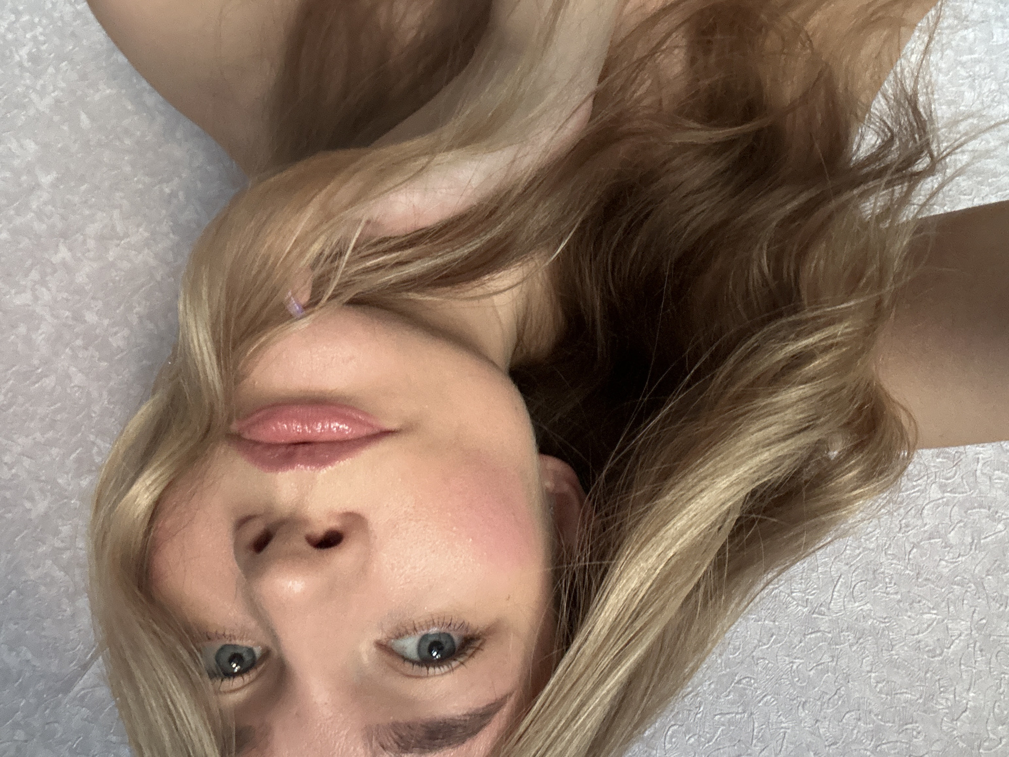 A young woman with blonde hair and blue eyes is shown in a close-up, angled shot. She is lying down, with her head tilted to the side. Her lips are painted with a glossy pink lipstick, and her cheeks are flushed pink, suggesting blush. Her right hand is raised and touching her hair, which is a mix of blonde and brown tones. The background is a textured, light-colored surface, possibly wallpaper or fabric. The image is lit from the front, casting soft shadows. The overall impression is of a casual, intimate portrait.