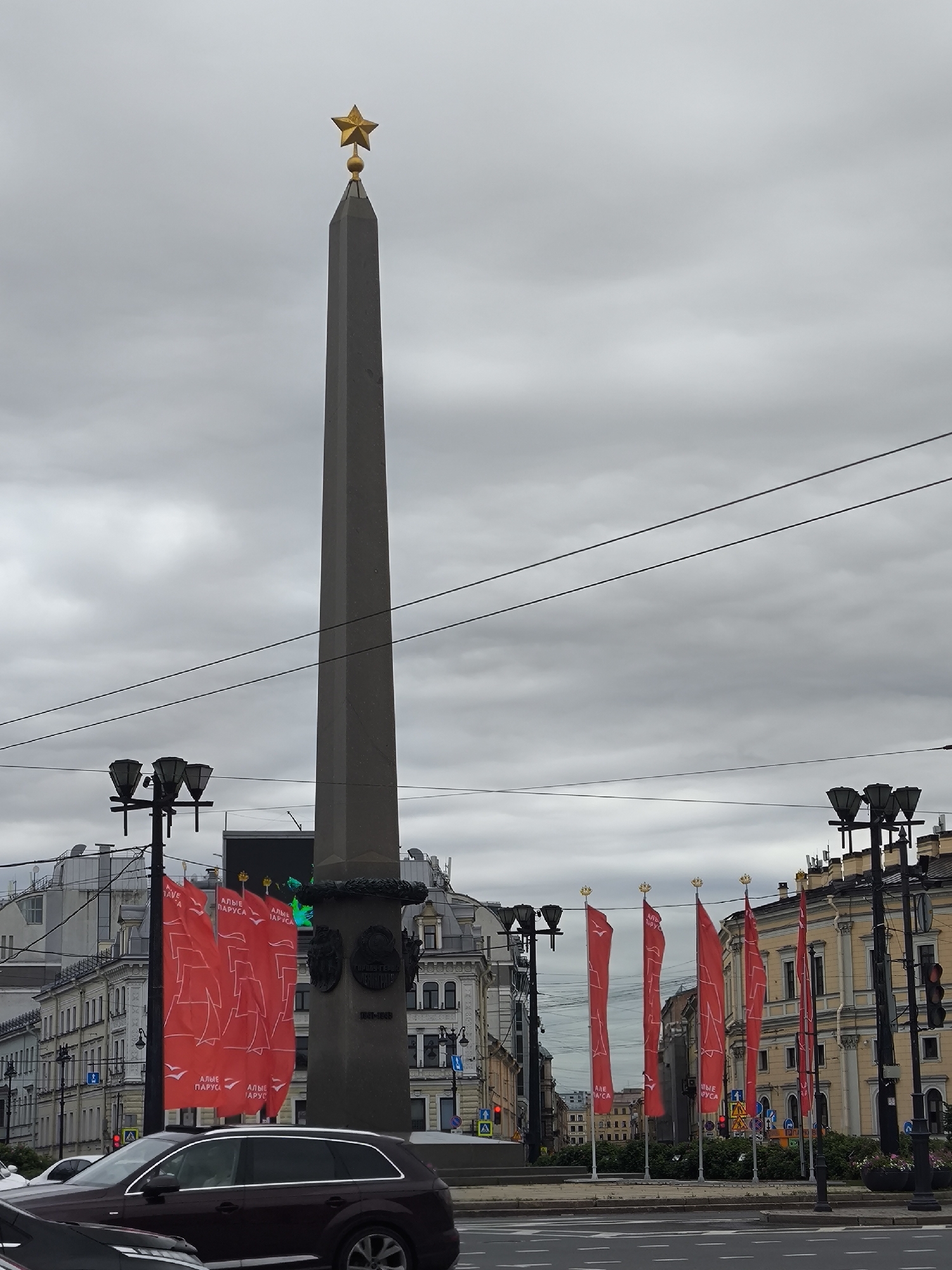 The image shows a view of the Rostral Columns on Strelka Spit in Saint Petersburg, Russia, on an overcast day. A tall, dark grey obelisk topped with a golden star stands prominently in the center of the frame. In front of the obelisk, several red flags with white sails and the inscription "АЛЫЕ ПАРУСА" (Scarlet Sails) are displayed. Several street lamps with multiple lights are visible on both sides of the scene. The background consists of multi-story buildings with classic architecture. A dark colored SUV is parked on the road in the foreground, with other vehicles partially visible. Traffic lights and street signs are also present. The sky is filled with heavy grey clouds.