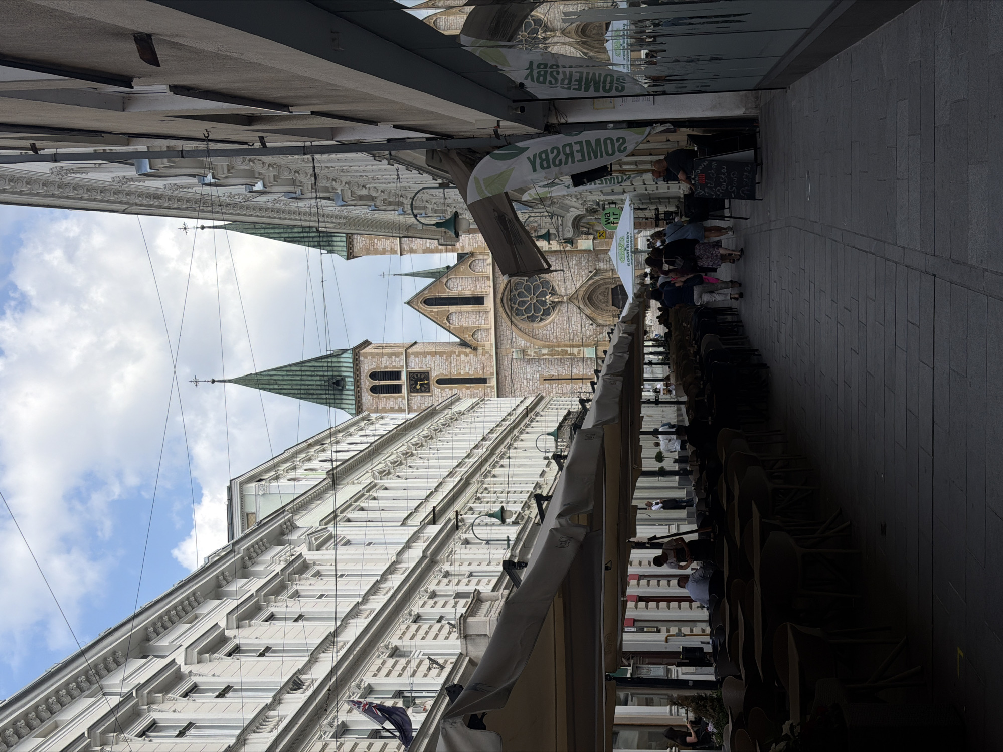 The image depicts a street scene in Sarajevo, Bosnia and Herzegovina, likely in the historic center. On the left, a long, multi-story building with ornate white facade and numerous windows lines the street. Overhanging awnings of a cafe extend across the sidewalk, shading rows of tables and chairs, some of which are occupied by people. In the background, a prominent stone cathedral with twin green spires and a large rose window is visible. The street is paved with stone and a number of people are walking along it. On the right side of the street, another building with a plain facade and windows is present, with advertisements and signage visible. The sky above is partly cloudy.