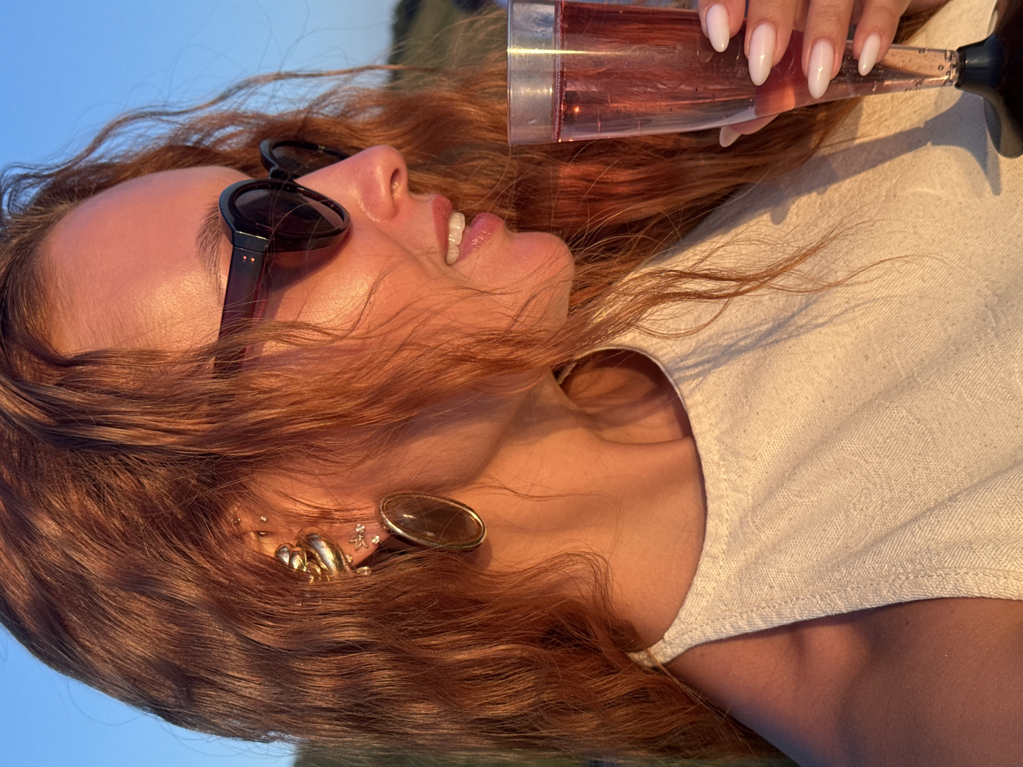 A woman with red hair, wearing sunglasses and a white top, is holding a glass of pink liquid. She has large gold earrings on her ears, and several smaller piercings above them. Her fingernails are painted a light pink color. She is outdoors, with a clear blue sky and a green, hilly landscape visible in the background. The lighting suggests it is late afternoon or early evening. The specific location in the city of Ольховка, Russia, cannot be determined from the visual information provided.
