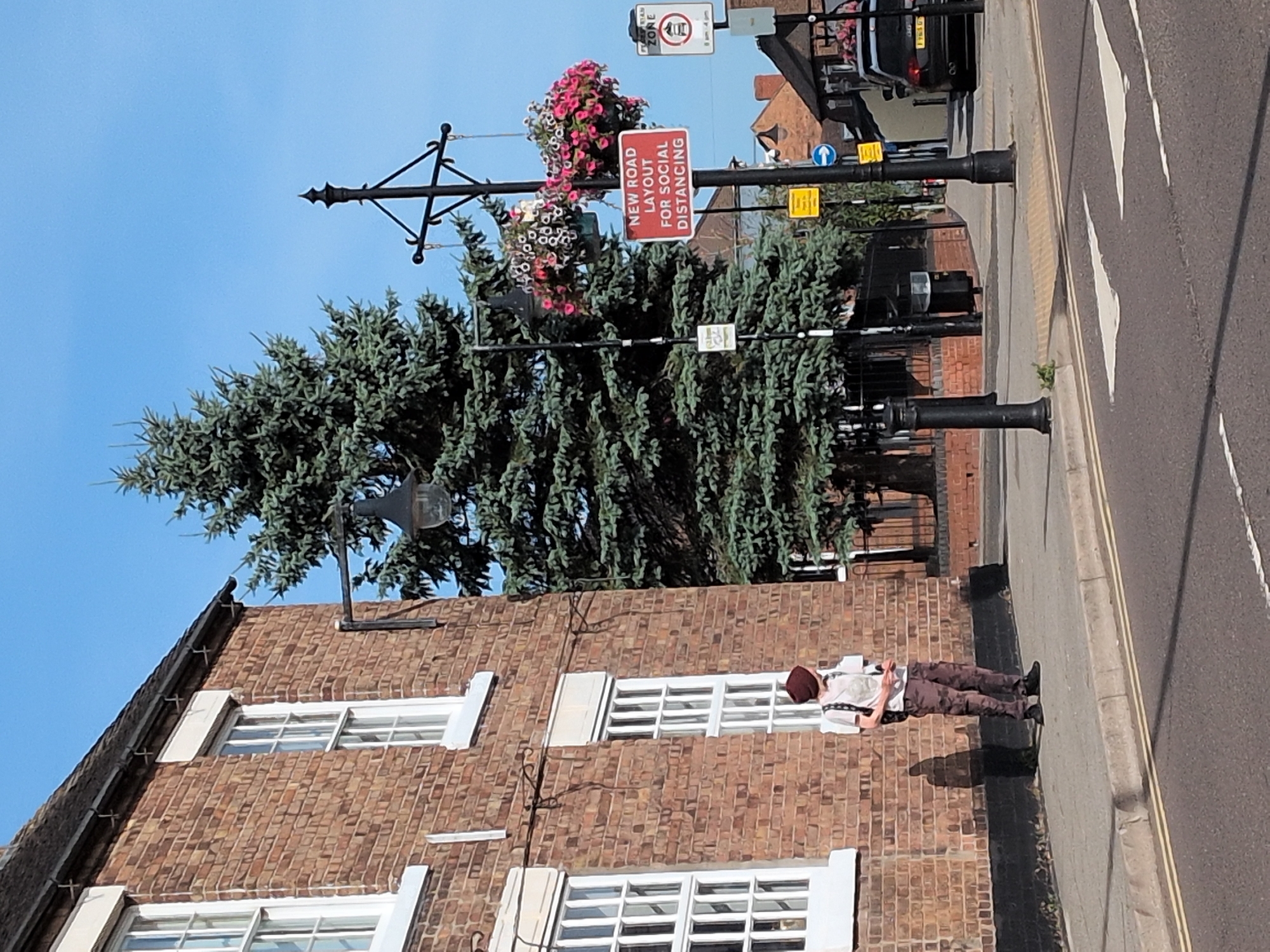 The image depicts a street scene in Dawley, United Kingdom, on a clear, sunny day. A person wearing a maroon beanie, a light-colored t-shirt, and camouflage-patterned trousers stands on a sidewalk next to a brick building. They are carrying a backpack and appear to be looking to the right. Adjacent to the building is a tall evergreen tree. A lamppost with hanging flower baskets stands on the sidewalk, adorned with a red sign that reads "NEW ROAD LAYOUT FOR SOCIAL DISTANCING." Another lamppost with a trash bin is visible further down the sidewalk. The street in the foreground features a pedestrian crossing with white stripes and yellow lines on the road. In the background, a black car with flowers on its roof is parked. The architecture suggests an older urban or town setting.