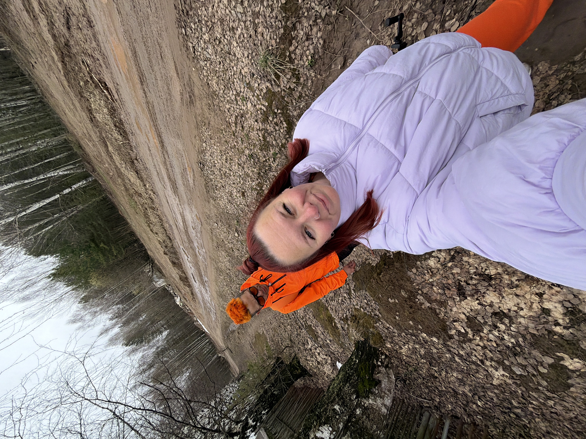 The image shows a woman with reddish hair and a light purple puffer jacket and orange pants lying on the ground in an outdoor setting. She is looking at the camera with a slight smile. Another person in an orange jacket is partially visible behind her. The ground is covered with dirt, leaves, and small pebbles. In the background, a dirt road runs alongside a forest of bare trees. Based on the location, this could be a wooded area or park near the city of Chaikovsky, Russia.