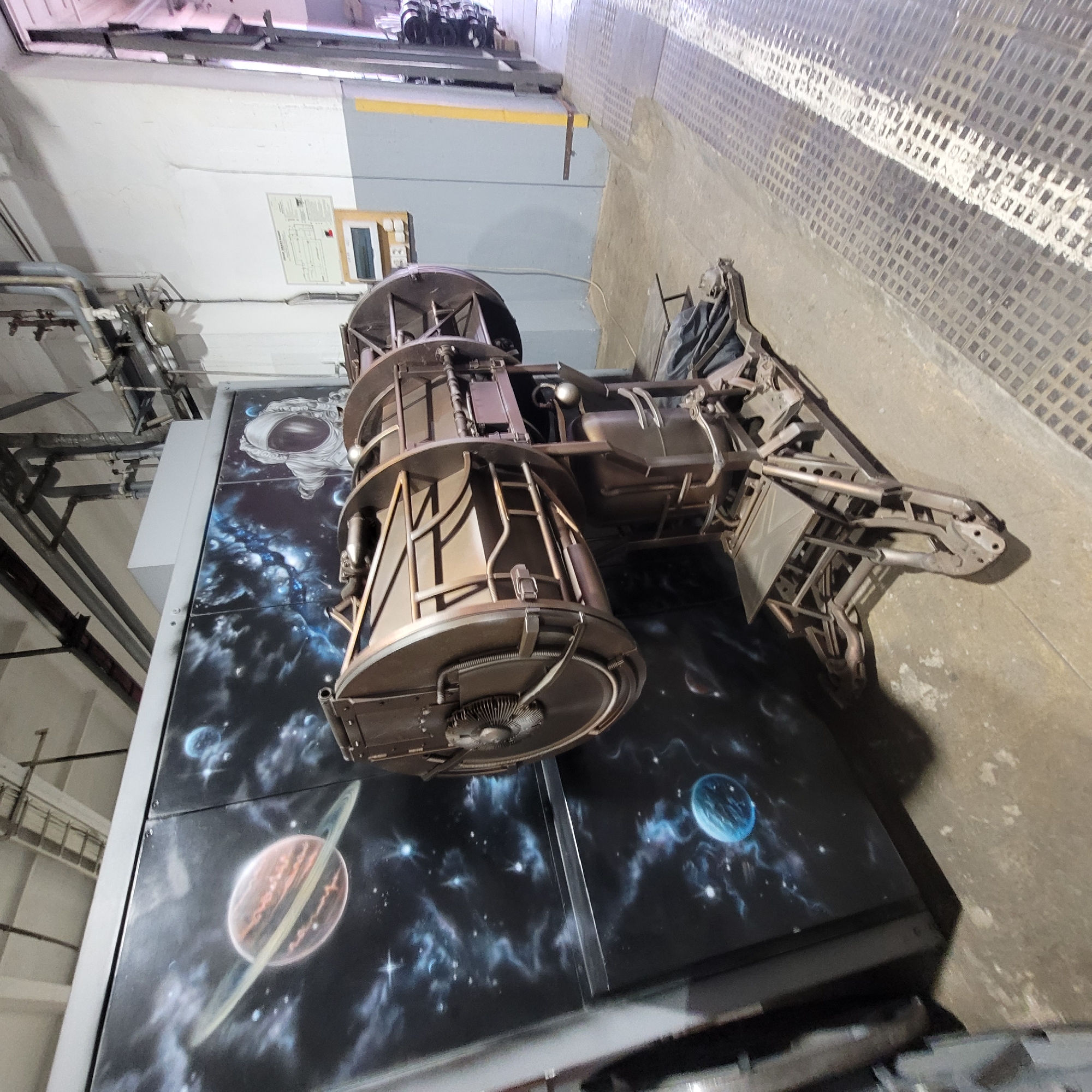 An indoor scene showcases a large, intricate metal structure resembling a futuristic machine, spacecraft, or industrial art installation. This object is positioned over a dark, vertical panel painted with a detailed space mural featuring planets, stars, and nebulae. One section of the mural prominently displays an astronaut's helmet.
The metal structure itself has a weathered, dark bronze or rust-colored appearance, composed of cylindrical sections, open frameworks, pipes, and various mechanical components. It features a fan-like circular element on one end and extends horizontally to a complex, skeletal form.
The setting appears to be an industrial or workshop environment, with white and grey walls, exposed pipes, conduits, and metallic rails visible in the background and overhead. The floor is concrete with a section of dark, square-patterned tiling. On a white wall, a document, possibly a technical diagram or schematic, is visible next to an electrical box or control panel, but its text is unreadable. The artificial lighting suggests an indoor location, possibly a museum, exhibition space, or production facility.