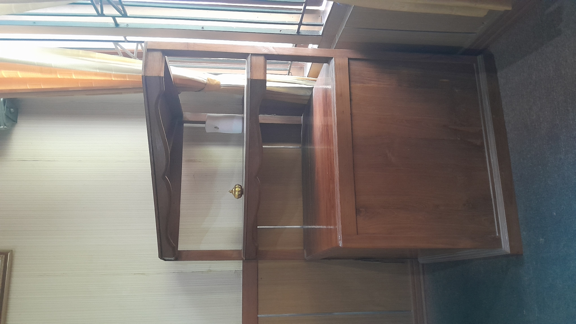 The image shows a wooden display shelf with two shelves, positioned against a wall with vertically striped wallpaper. The shelf unit is made of dark wood. On the top shelf, there is a small, golden decorative object and a white cylindrical object. The lower part of the shelf unit is a solid wooden cabinet with a large drawer or door front. The floor is covered with dark carpet. To the right of the shelf unit, there is a window with light-colored curtains and a view of what appears to be a barred window. The overall impression is of an indoor setting, likely a room in a residential or commercial space in Blora, Indonesia. No people are visible in the image.