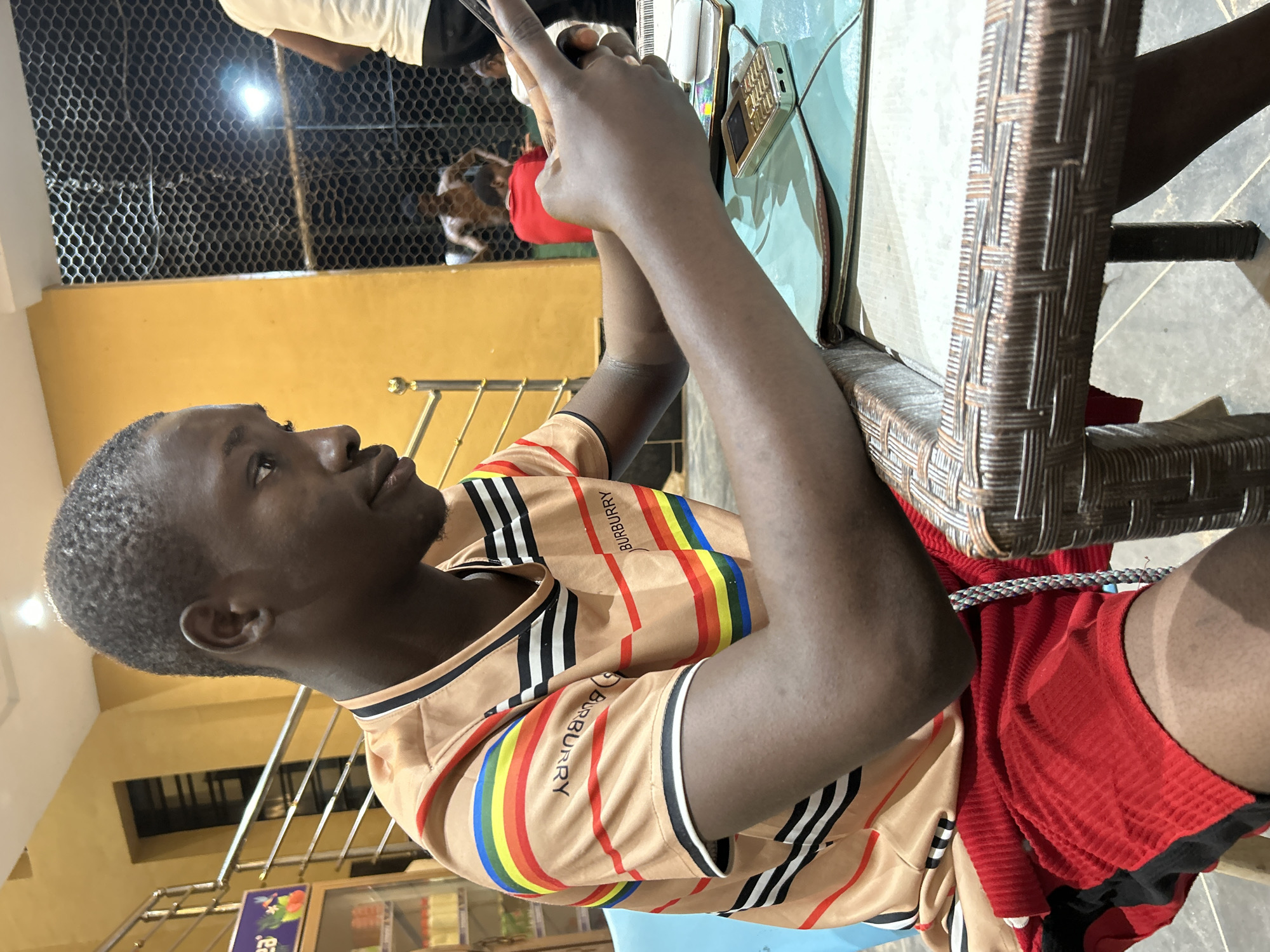 A young Black male with short hair is seated at a table, looking at a mobile phone in his right hand. He is wearing a tan t-shirt with horizontal stripes of black, white, and rainbow colors, and red shorts with black trim. The table has a woven edge and a turquoise mat on top. A small gold mobile phone and a white notepad are on the table. In the background, a metal railing and a yellow wall are visible, along with a wire mesh fence. Behind the fence, several other people are present, some of whom appear to be playing a game. The setting appears to be an outdoor or semi-outdoor location with artificial lighting. The image is taken from a medium-close perspective, focusing on the young male.