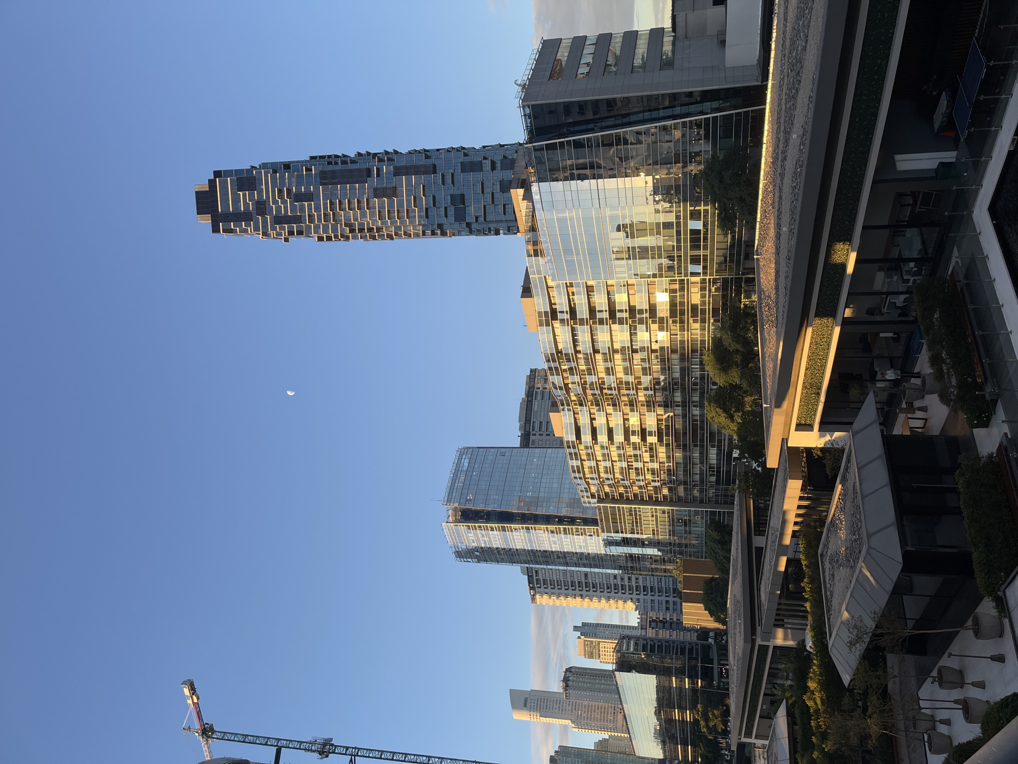 The image shows a cityscape in Buenos Aires, Argentina, featuring several modern high-rise buildings with glass facades reflecting sunlight. A distinct, slender tower with a staggered, irregular pattern is prominent on the left. A construction crane is visible in the lower left corner. A crescent moon is faintly visible in the clear blue sky. The scene appears to be taken from an elevated position, possibly a rooftop. Part of a building with greenery and outdoor seating is visible in the foreground on the right.