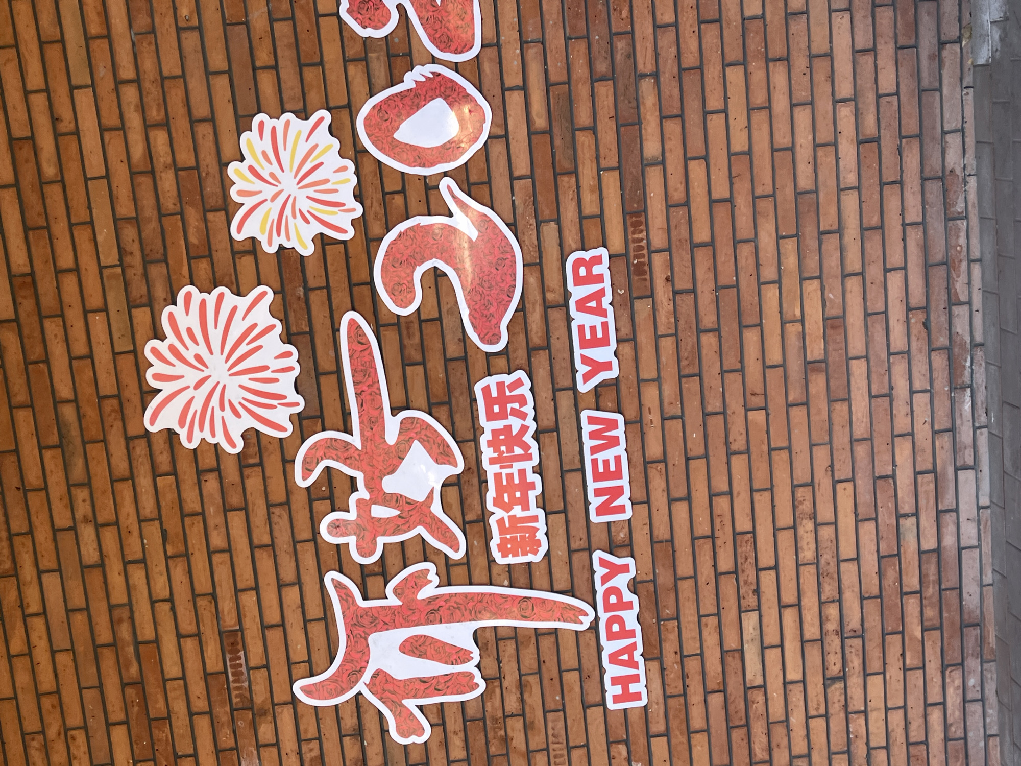 The image shows a brick wall with celebratory decorations. The decorations include paper cutouts of fireworks, the Chinese characters for "Happy New Year," the English phrase "Happy New Year," and the year "2023." The decorations appear to be placed on the brick wall, suggesting it's part of a celebratory display for the Lunar New Year. Given the writing on the wall, it could be a shop or residence wall. It is likely located in Changchun City, China, given the prompt.