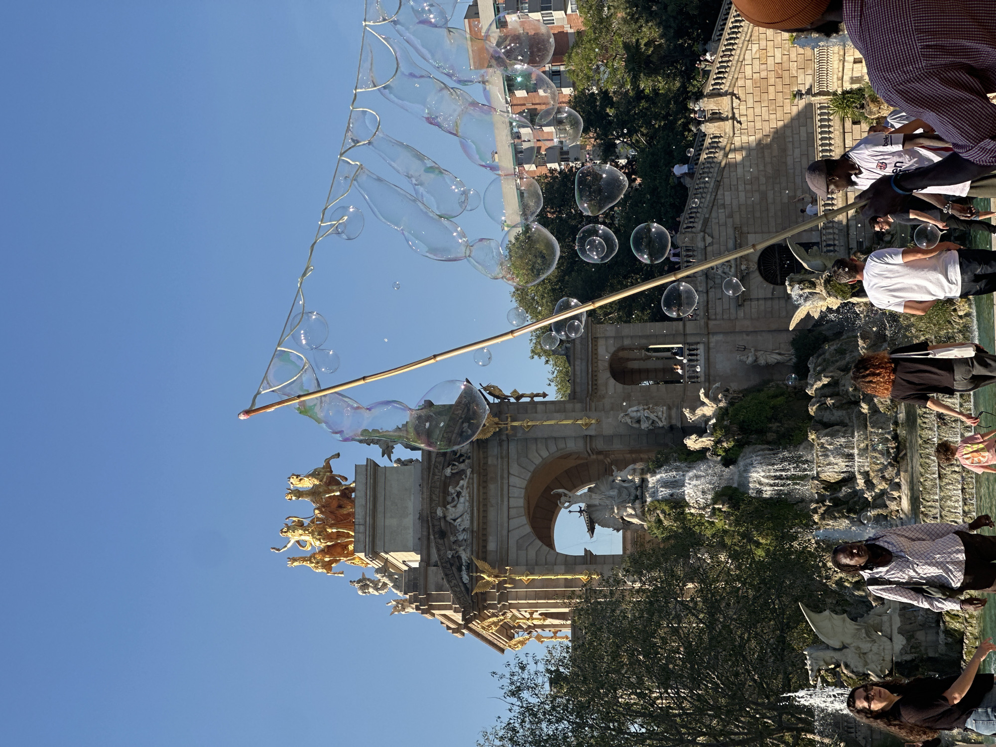 This image depicts the Arc de Triomf in Barcelona, Spain, under a clear blue sky. A device for creating large bubbles is being used, resulting in numerous bubbles floating in the air. The arc's architectural details, including golden statues, are visible. The surroundings include trees and a stone structure with a waterfall feature. People are present, standing near the water and observing the bubble-making activity. Some are on steps leading up to the structure.