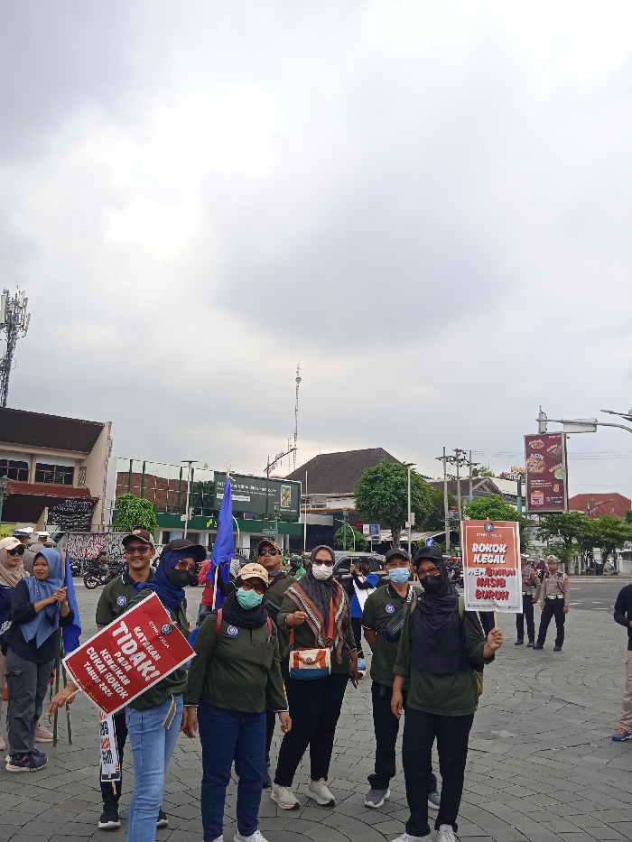 The scene is located in Yogyakarta, Indonesia at coordinates -7.783 latitude and 110.367 longitude. It depicts a group of nine individuals standing on a paved street with a few buildings in the background. The individuals are mostly facing the camera and are holding banners with Indonesian text. Seven of the individuals are wearing face masks. The individuals appear to be engaged in a demonstration or protest. One of the individuals is holding a banner that states "TIDAK!" with text below that. The other individual is holding a banner that states "ROKOK ILEGAL MERUGIKAN MASYARAKAT BURUH". There are also a few other people standing further back in the background, some of them appearing to be observing the group. There is a large banner behind the group with Indonesian text. It is a cloudy day.