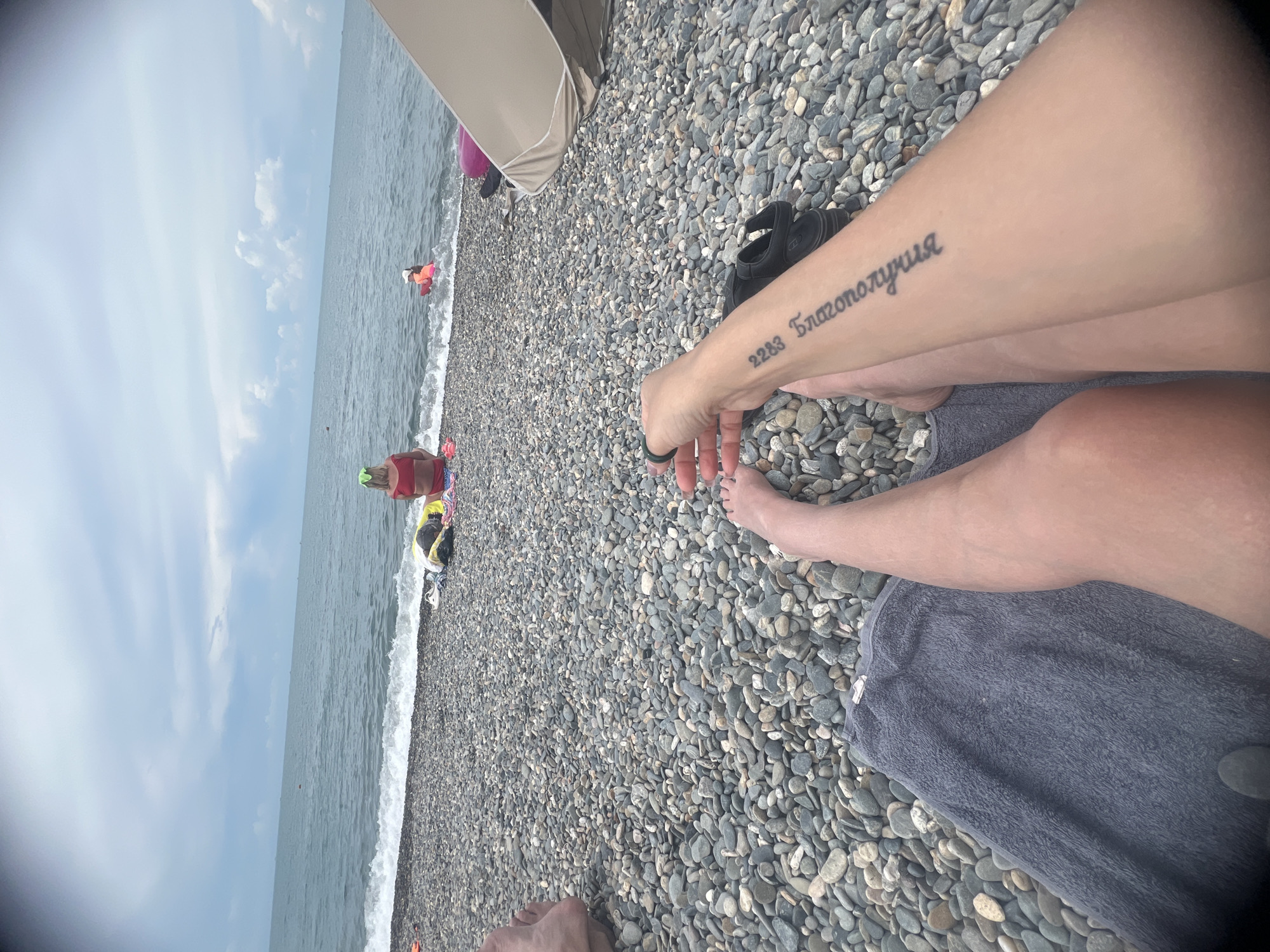 The image shows a first-person perspective of a person relaxing on a pebble beach. The person's legs and arm are visible in the foreground, with a tattoo on the arm. A grey towel is laid out on the pebbles. In the background, the sea is visible with waves breaking on the shore. Several other people are present on the beach, including a woman in a red bikini and a child in a pink floatation device in the water. A beach umbrella or tent is partially visible on the right side of the frame. The sky is overcast with some clouds.