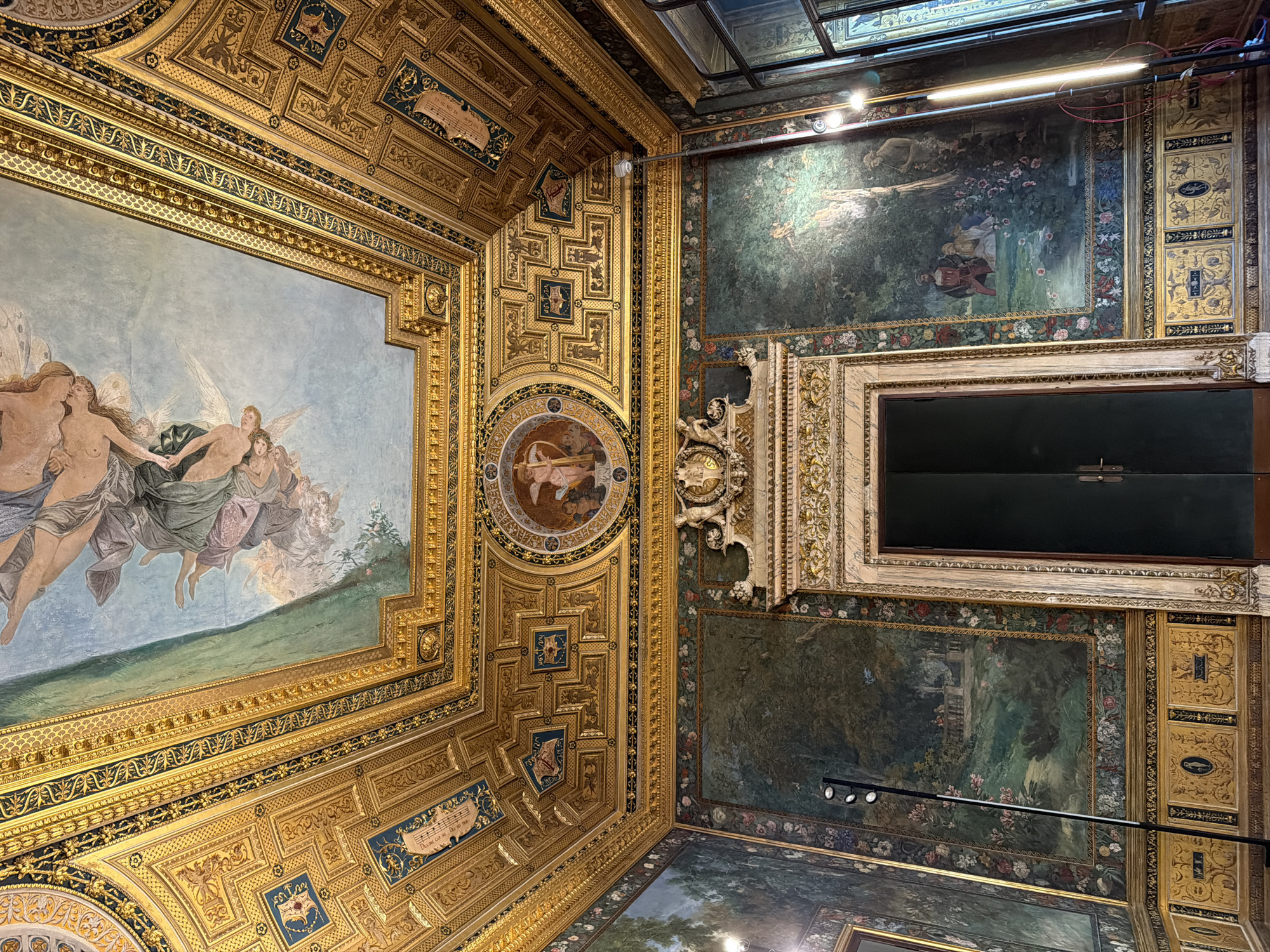 Captured from a low-angle perspective, the photograph showcases the elaborate ceiling and wall art of a room. On the left, a large painting framed in gold depicts figures in flight against a light blue sky. The ceiling is adorned with intricate gold detailing, interspersed with smaller paintings and designs. To the right, the walls are covered in darker, nature-themed paintings with floral borders. A doorway or window is framed in ornate marble or light-colored stone. The lighting is a mix of natural light coming through the skylight and artificial light from fixtures. The location is in the city of Unknown, Unknown.