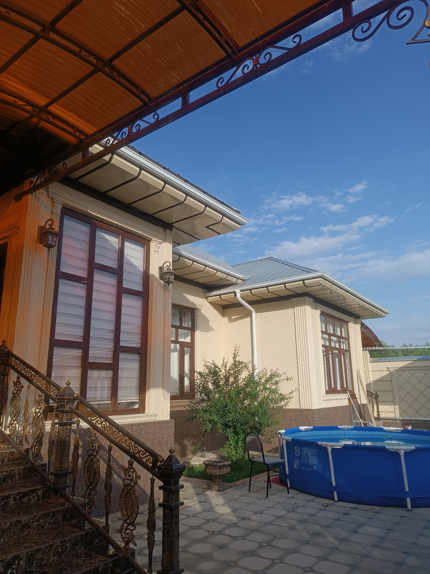 The image depicts a sunny outdoor scene in the city of Myrza-Ake, Kyrgyzstan. In the foreground, a set of dark marble stairs with an ornate wrought iron railing leads up to a light-colored building. The building features large, dark-framed windows with blinds and decorative lamps on the exterior walls. Above the building, a brown corrugated metal awning extends outwards.
To the right of the building, a bright blue inflatable swimming pool is visible on a patterned paved patio. A black metal chair sits next to the pool, and a small green bush is planted nearby. The background shows more of the building, a fence, and a clear blue sky with scattered white clouds. The overall impression is one of a residential property on a clear day.