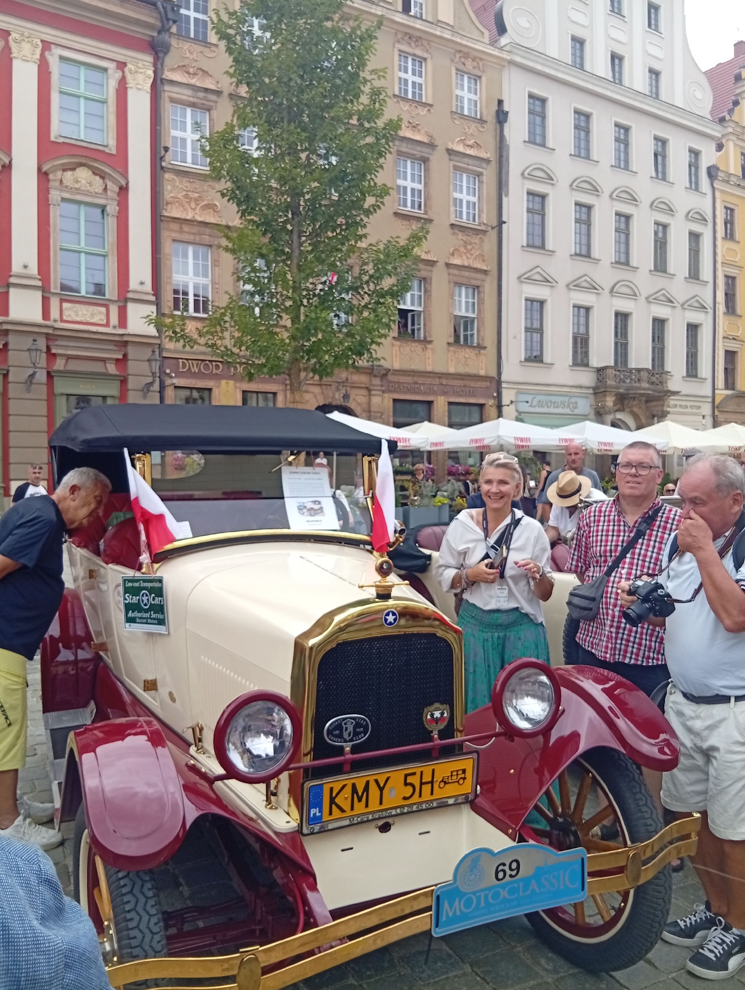 This image shows a group of people gathered around a vintage cream and maroon colored car in a city square in Wrocław, Poland. The car has a black canvas roof and prominent gold trim. It features two flags, one red and white, and a license plate reading "KMY 5H". There is also a blue sign with "69 MOTOCLASSIC" printed on it.
In the foreground, a man in a dark t-shirt and yellow shorts leans towards the car. To his right, a woman in a white shirt and green skirt smiles, holding a phone or camera. Next to her, a man in a red and white checkered shirt holds a camera. To the far right, another man in a white shirt and light-colored shorts stands, touching his face. In the background, several buildings with European architecture line the square, some featuring awnings and signs for businesses, one of which reads "Laworska." People are visible further in the background, and trees add greenery to the urban setting. The ground is paved with cobblestones.