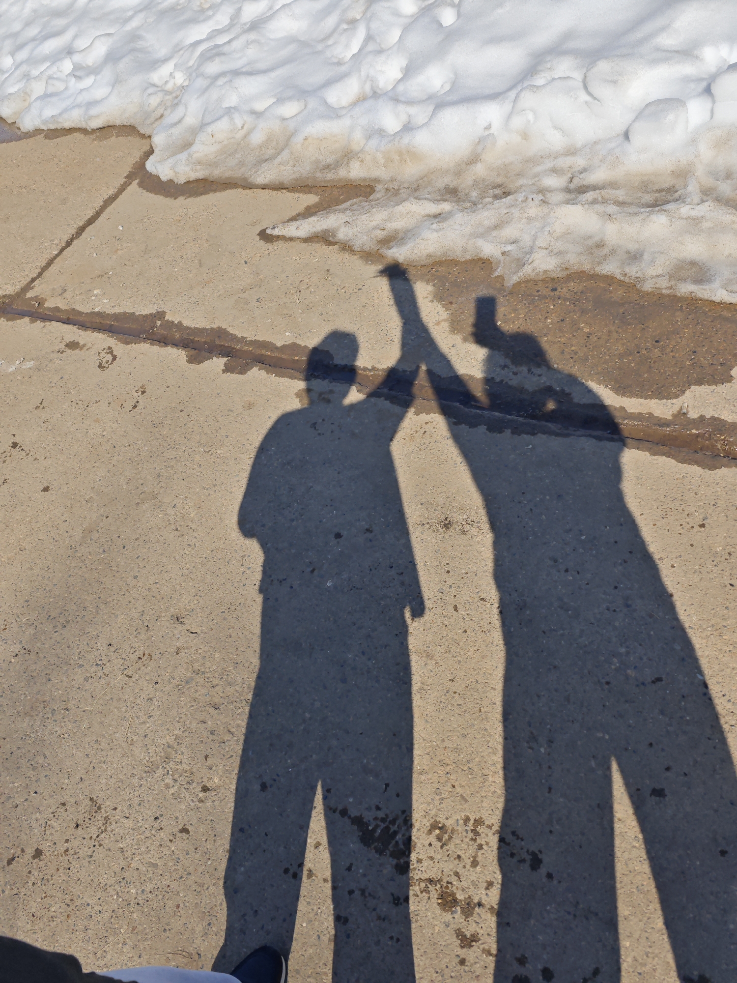 The image shows two shadows of individuals appearing to high-five each other, cast on a concrete surface. Adjacent to the concrete, there is melting snow. The location is assumed to be in or near Марковский, Russia. The individuals casting the shadows are not visible.