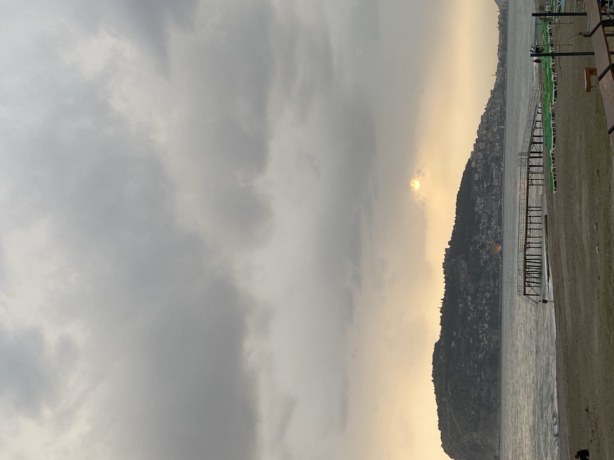 The image shows a beach scene in Alanya, Turkey. The sky is overcast with heavy, gray clouds. A faint yellow sun is partially visible through the clouds. A coastline with dense urban development is seen to the right, rising up a hillside. The shoreline is sandy, transitioning into water. A pier or wooden structure extends into the water. Chairs and possibly other beach furniture are present on the sand. The photograph is oriented sideways, with the horizon running vertically through the frame.