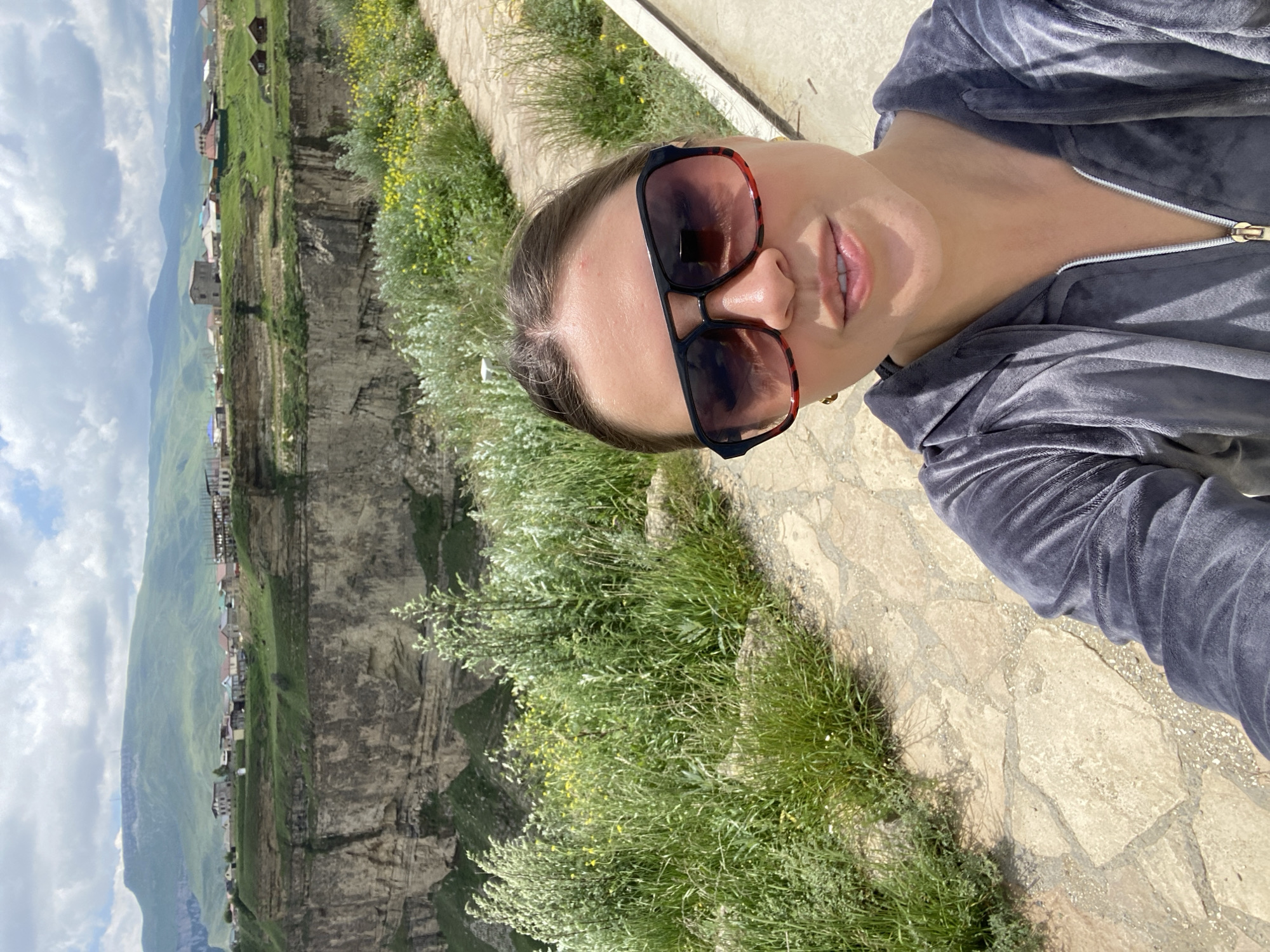 A woman is taking a selfie outdoors. She is wearing sunglasses and a grey hooded jacket. The background shows a village on the edge of a canyon with mountains in the distance. The location is the Tsolotlinsky Canyon, near the village of Khunzakh in Dagestan, Russia. The canyon is a popular tourist destination known for its dramatic cliffs and scenic views.