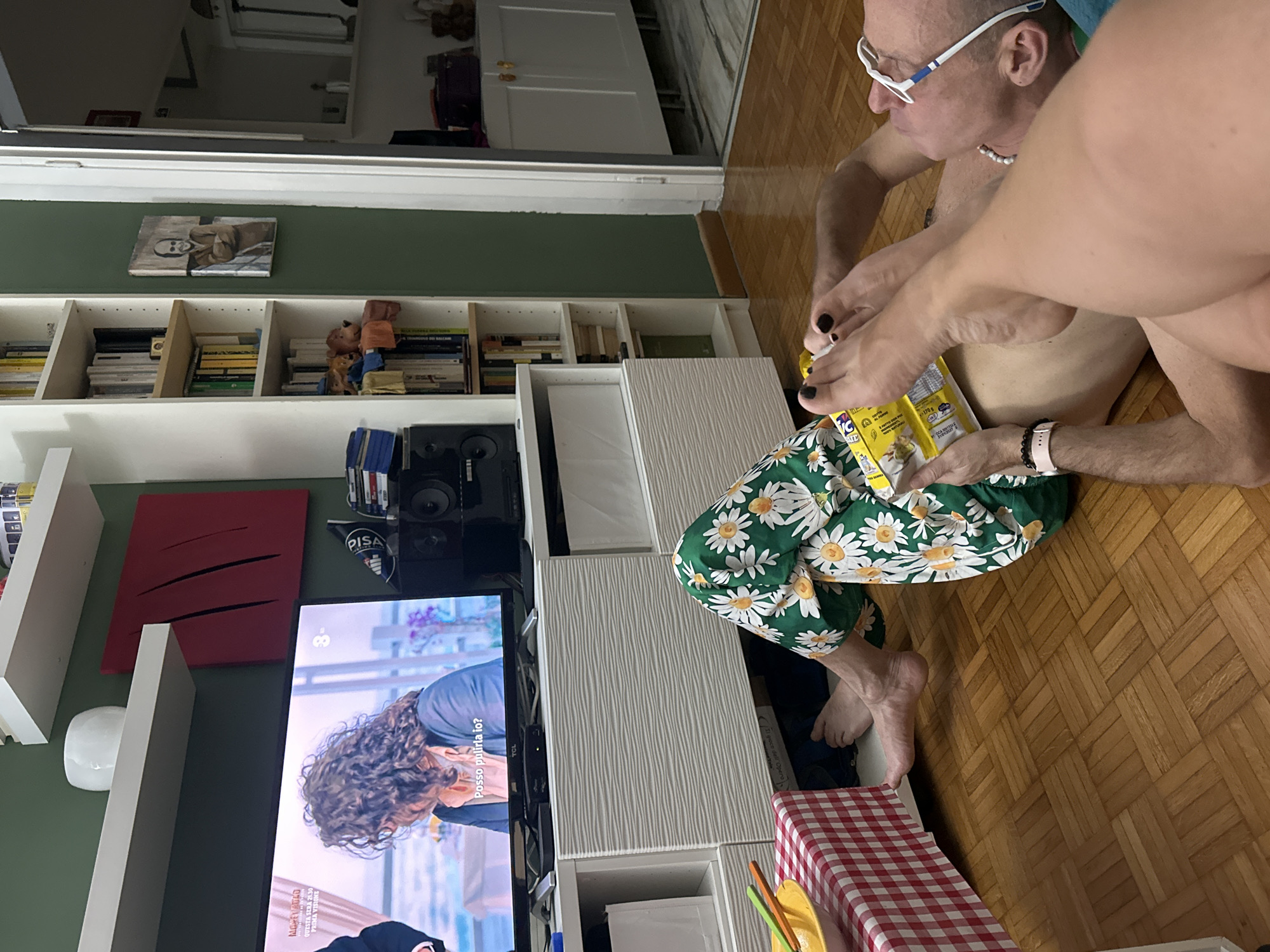 The image depicts a living room scene with a man lying on a wooden floor, propped up by a cushion. He is wearing a pearl necklace and glasses, and has his legs extended. Another person's legs, clad in floral-patterned pajama pants, are resting on his legs. The man is holding a box of cereal, and his toes are adorned with black nail polish. In the background, a television is on, displaying a scene with a woman with curly hair. To the left of the television, a white cabinet unit is visible, with a red and white checkered tablecloth laid out on top of it. Shelving units filled with books and decorative items line the wall behind the television and to the right of the scene. A white door with a dark frame is also visible on the right side of the image, leading to another room. The overall setting suggests a relaxed domestic environment. There is no indication that the location is Milan, Italy.