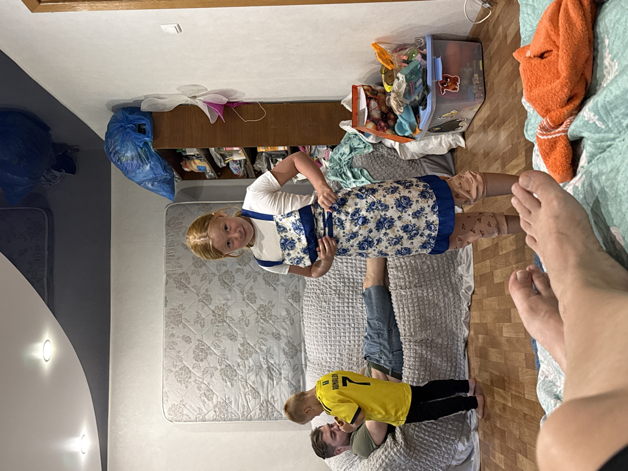 The image shows a family in an indoor living space. In the foreground, a person's legs and feet are visible, lying on a turquoise blanket. Further back, a young girl with blonde hair is standing and wearing a white t-shirt and a blue and white floral apron. To her left, a young boy in a yellow t-shirt with "RONALDO 7" printed on it is facing a man lying on a textured grey couch. The man is wearing a green t-shirt and blue jeans. The room has wooden flooring and white walls. A dark grey ceiling with recessed lighting is visible. A large mattress with a floral pattern leans against the wall behind the couch. To the right, a wooden shelf unit is filled with various items, including blue plastic bags and other assorted objects. In front of the shelves, a clear plastic storage bin filled with toys and other items is on the floor. A bright orange towel is also visible on the turquoise blanket in the foreground. The overall impression is a casual, domestic scene. The image is identified as being taken in the city of Valday, Russia.