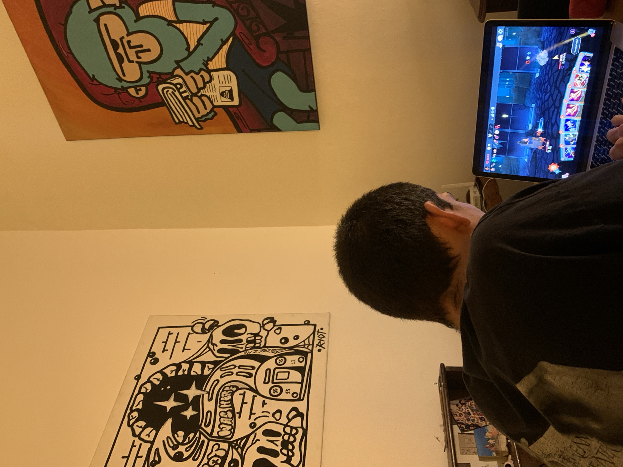 A person with short, dark hair is sitting in front of a laptop, seemingly playing a game. The laptop is positioned on a desk, showing a game interface with colorful characters and symbols. The person is wearing a dark-colored shirt. On the wall behind the person, there are two pieces of art. The upper one is a cartoon-style image featuring a character with green skin and orange surroundings. The lower art piece is a black and white drawing with abstract designs and patterns, including skull-like figures and writing. There are other items on a lower shelf or cabinet to the right of the person. The image is taken indoors under artificial lighting. The location cannot be determined based solely on the visual information.
