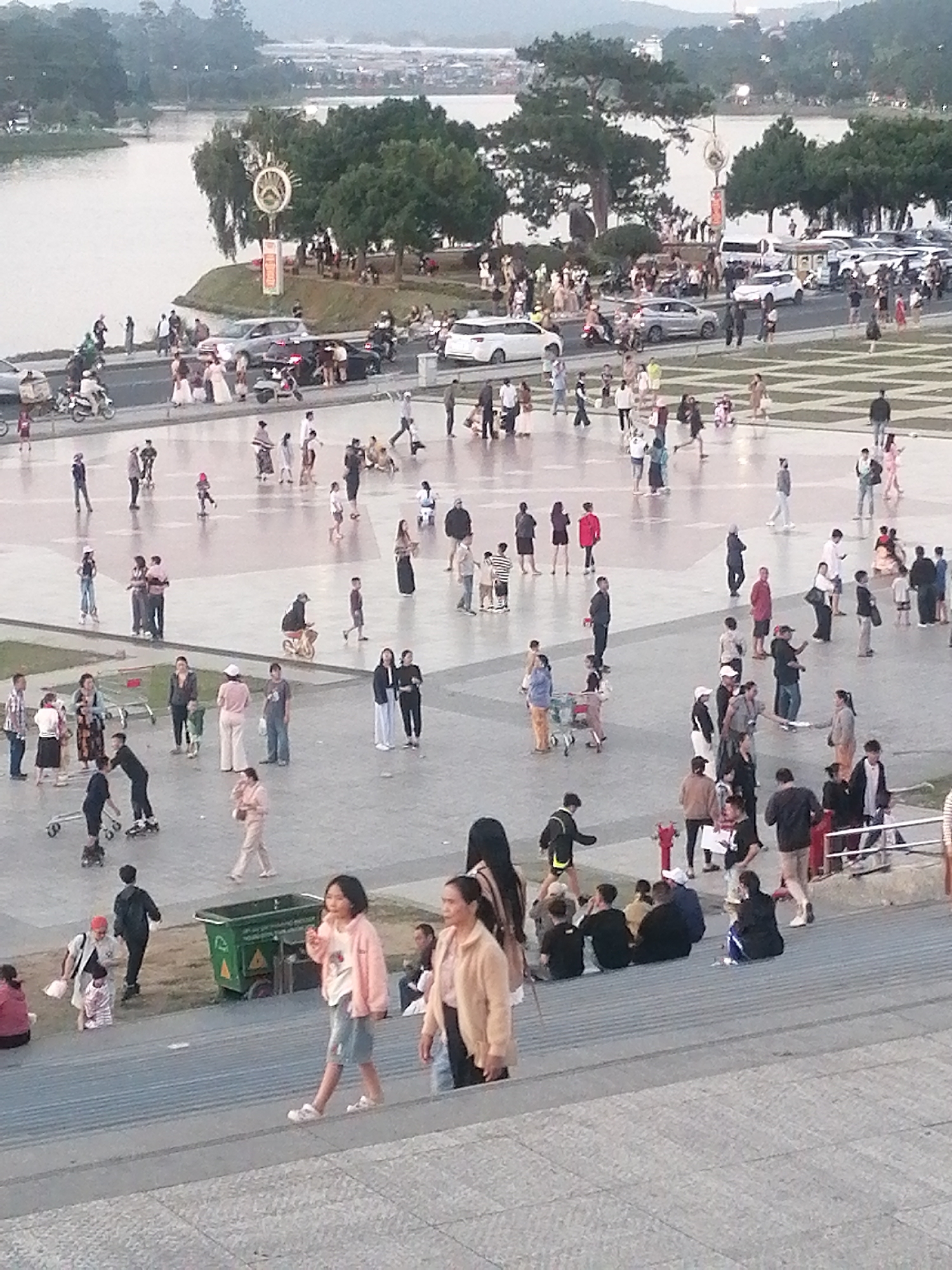 The image depicts a panoramic view of Xuan Huong Lake in Da Lat, Vietnam, on an overcast day. The foreground shows a wide paved area with a checkered pattern, where numerous people are gathered, engaged in various activities. Some are walking, others are sitting, and a few are on roller skates. A green waste bin is visible on the left side of the paved area. In the mid-ground, there is a road with cars, motorcycles, and more pedestrians. The background features a lush green hillside dotted with trees, and beyond that, a body of water, presumably Xuan Huong Lake. The distant landscape is characterized by hills and buildings, suggesting an urban setting. The overall atmosphere appears to be one of a leisurely outing in a public space, with a mix of locals and possibly tourists.