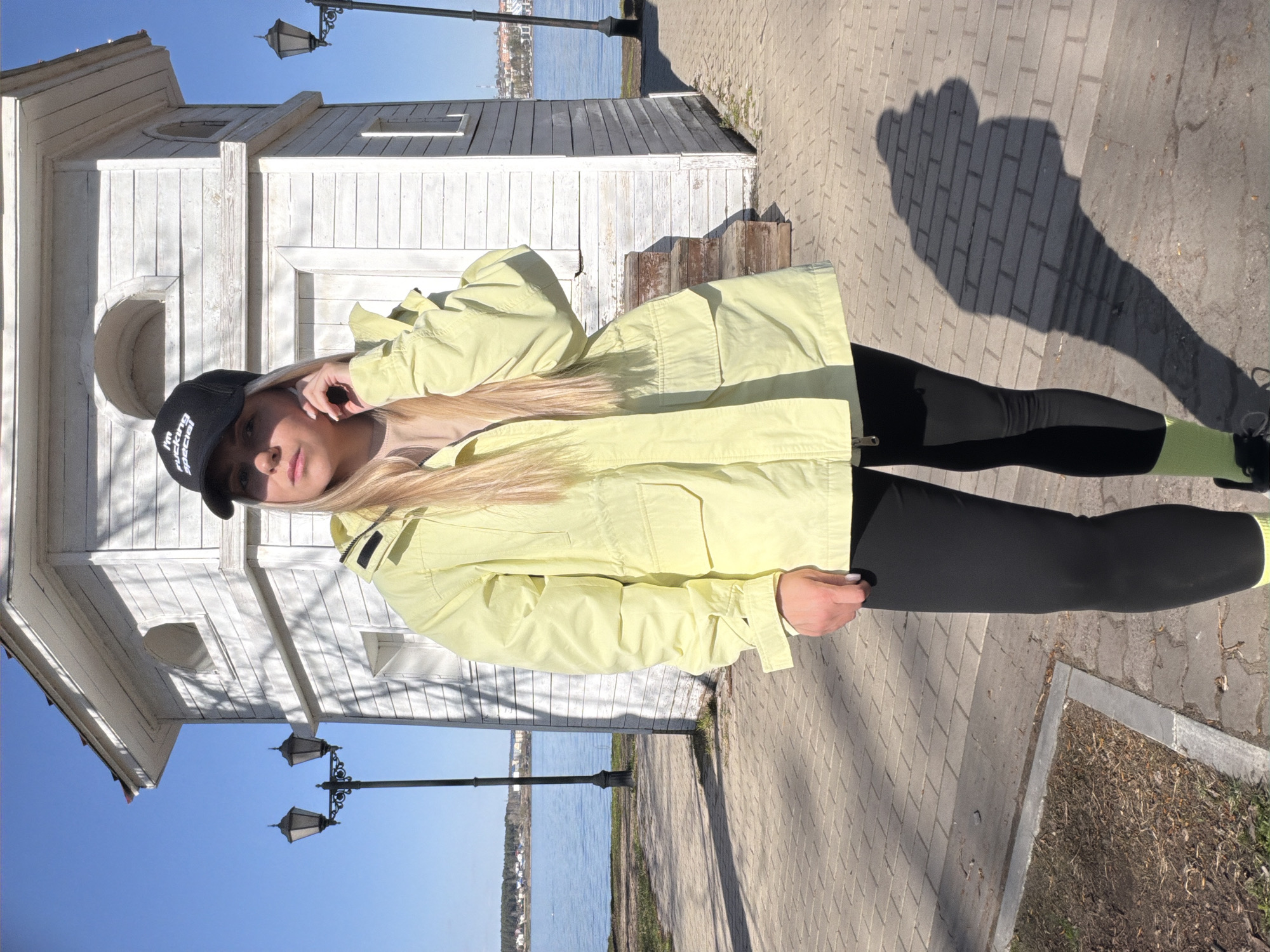 A blonde woman wearing a black baseball cap, light yellow jacket, and black leggings stands on a brick-paved walkway. She is standing near a small, white wooden structure, potentially a building or decorative gazebo. Behind her, there is a body of water, with visible structures across the water. Lamp posts are visible near the woman and along the walkway. The weather appears sunny, as indicated by the shadow cast on the ground. The location is likely Votkinsk, Russia due to the architecture and landscape characteristics.