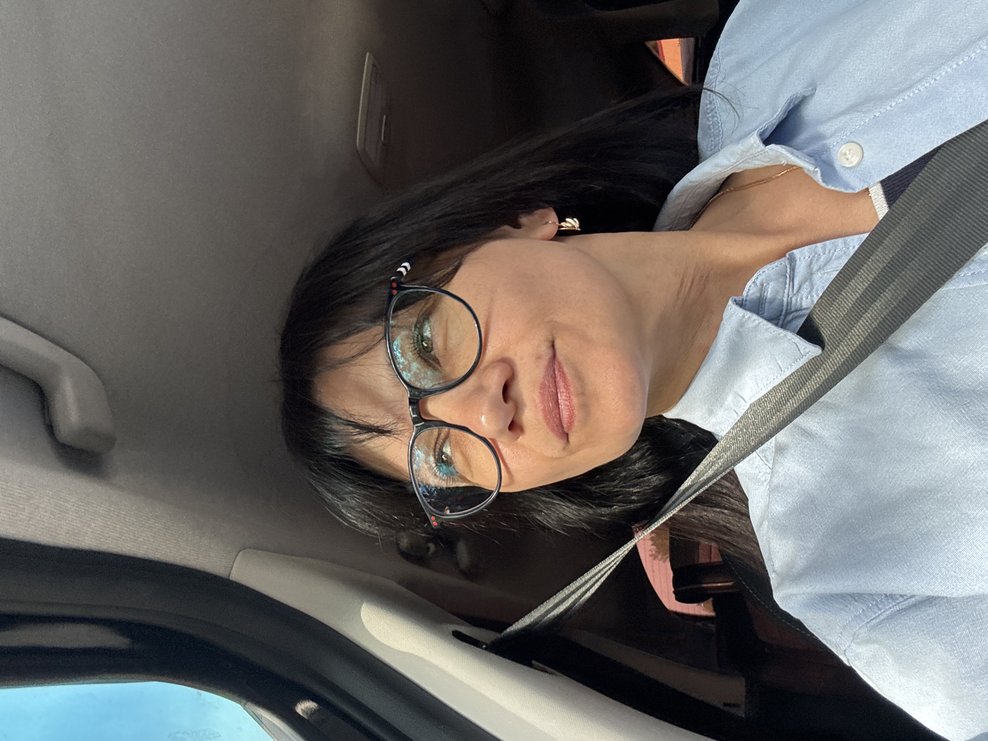 A person wearing glasses and a blue shirt with a seatbelt on is in a car. The location is unknown.