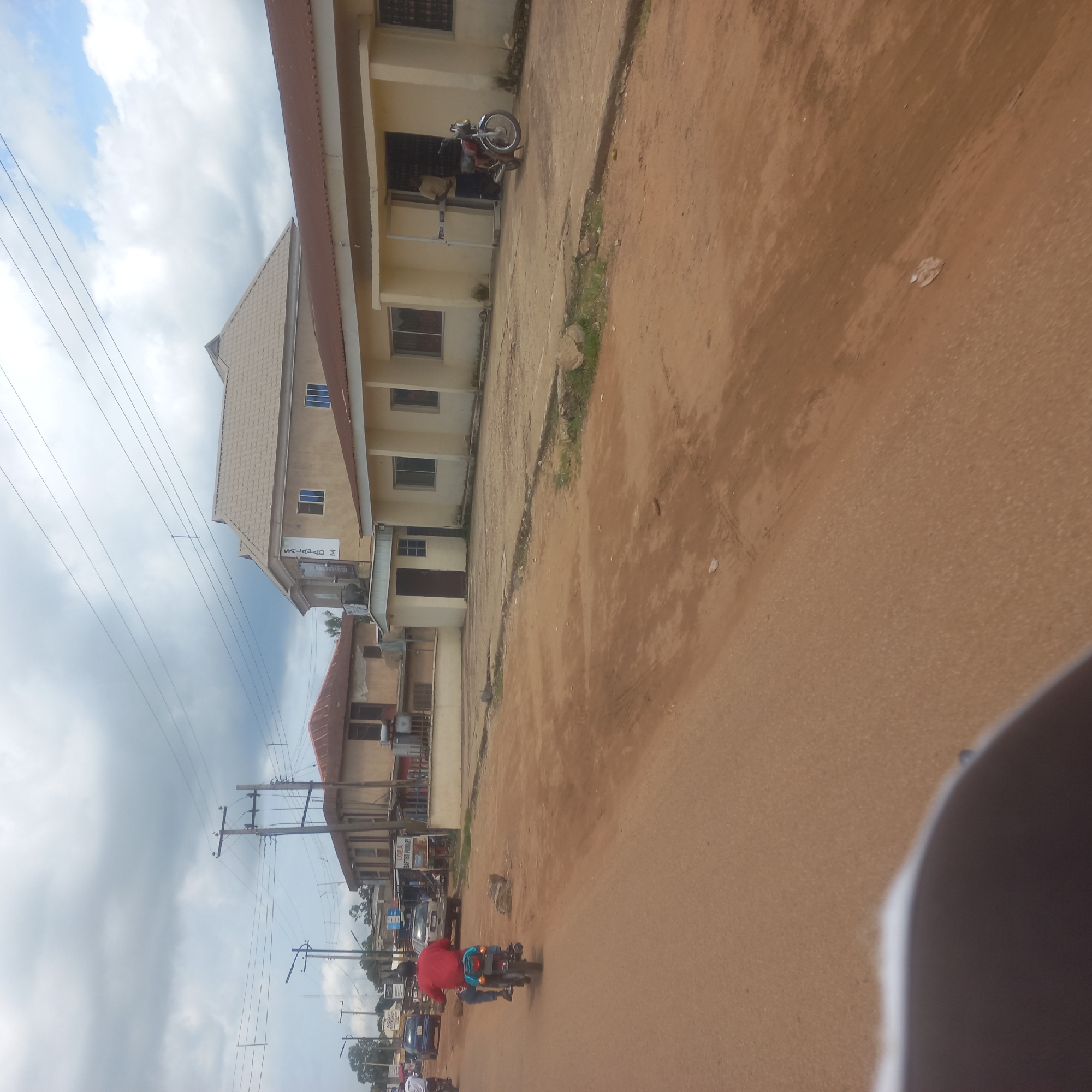 In Mangu Town, Nigeria, this image captures a street scene under a cloudy sky. The foreground shows a dusty, reddish-brown dirt road. On the left side of the road, a person wearing a red shirt is riding a motorcycle. Further down the road, several buildings are visible. To the right, a light-colored, single-story building with a red-brown roof has several windows and an entrance where a person in a light-colored shirt is standing near a motorcycle. Adjacent to this building is another structure with a similar roof and multiple windows. Behind these buildings, another building with a lighter roof and a darker upper section is visible. Power lines crisscross the sky above the buildings. A blue car is parked on the left side of the road in front of a building with a sign that reads "NOOZI BOOKSHOP". Another car is parked further down the road near a building with a sign that says "LGEA BAPTIST PRIMARY".