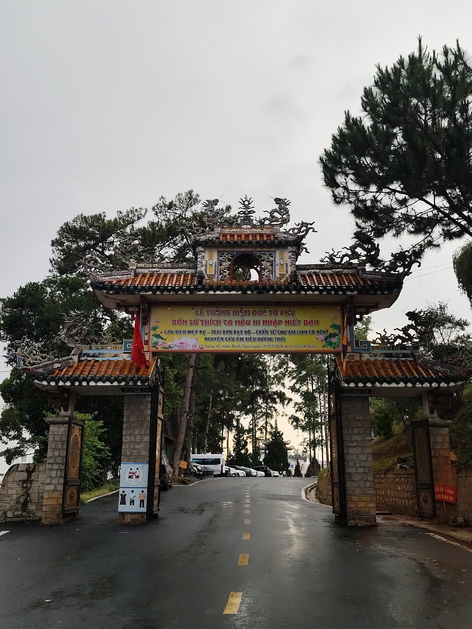 The image captures the entrance to the Truc Lam Zen Monastery in Da Lat, Vietnam. The gateway is a traditional Vietnamese architectural structure with an ornate tiled roof, stone pillars, and intricate decorations. A banner is hung across the top, displaying Vietnamese text related to a Buddhist ceremony. The sky is overcast, and the road leading through the gate is wet, reflecting the light. In the background, trees line the road, with cars parked along the side. A sign is visible on the left pillar, depicting icons of people and their behavior requirements, while a warning sign written in Vietnamese is on the right pillar.