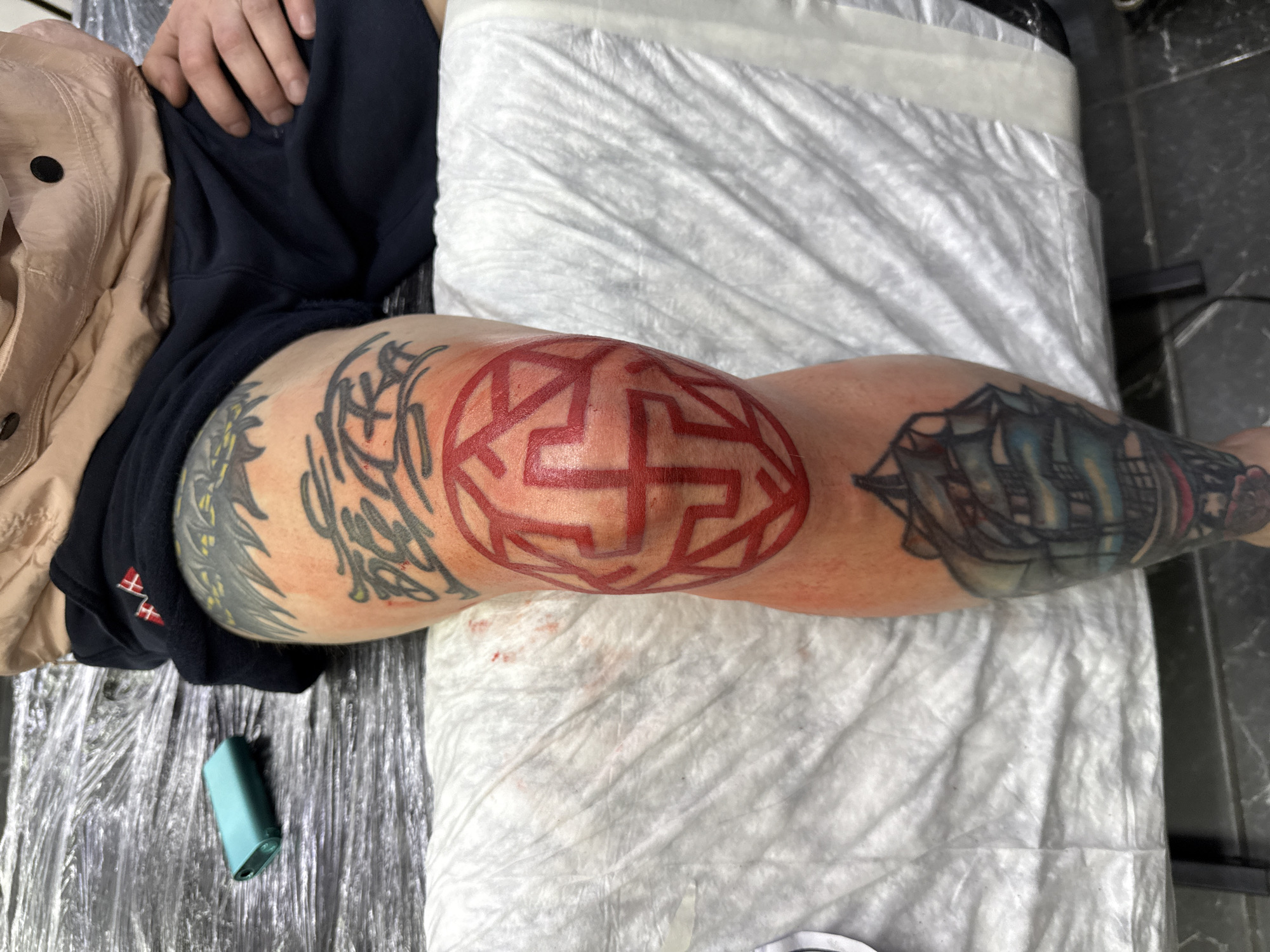 An overhead shot captures a person's lower body, focusing on a leg adorned with multiple tattoos, presumably in a tattoo studio in Moscow, Russia. The individual is lying on a surface covered with white disposable medical paper and plastic sheeting. A dark blue garment, possibly shorts or sweatpants, and a light brown jacket are visible on the upper left. A hand rests on the dark fabric at the top of the frame.
The central focus is a new, large red tattoo on the knee, featuring a complex, geometric, and maze-like circular design that appears fresh, indicated by the surrounding redness on the skin. To the upper thigh, a black and grey tattoo with abstract or tribal-like patterns is partially visible. Further down the leg, on the calf area, another existing color tattoo depicts a sailing ship or galleon with intricate rigging.
On the plastic-covered surface next to the leg, a small, turquoise or dark green rectangular electronic device, possibly a vape or charger, is placed. The background shows a dark, possibly tiled or concrete floor, suggesting an indoor, professional setting. A small red and white emblem, appearing as two stylized vertical bars connected by a horizontal one, is visible on the dark blue fabric of the shorts near the hip. The lighting is bright and even, typical of an indoor studio environment.