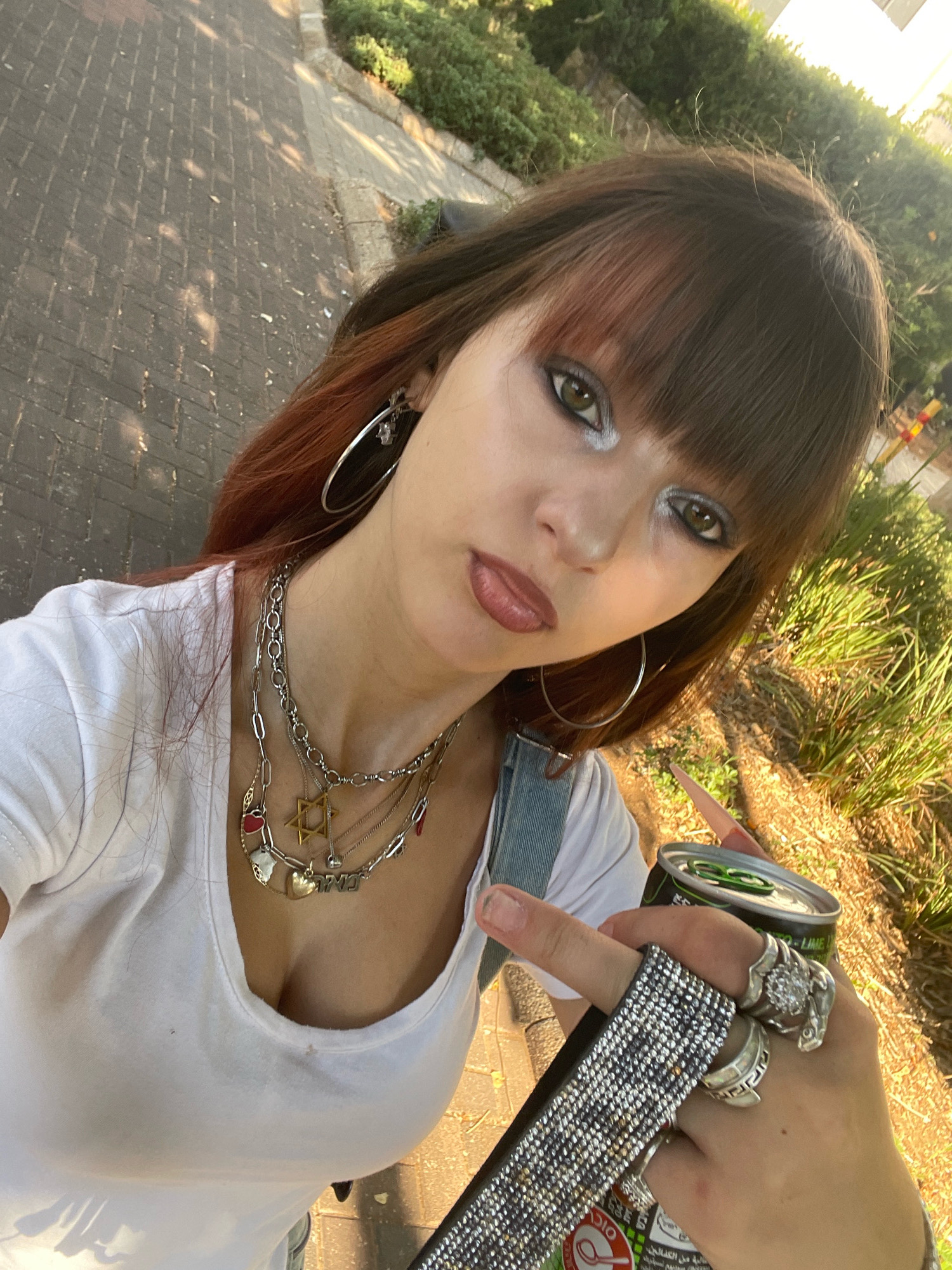 A young woman with long brown hair and bangs takes a selfie outdoors in Raanana, Israel. She is wearing a white t-shirt and multiple silver necklaces, including a Star of David pendant. Her makeup includes bold silver eyeshadow and dark lipstick. She is holding a can of an energy drink and a phone with a bedazzled case. In the background, there is a paved walkway, lush green bushes, and a building. The sunlight suggests it is daytime.