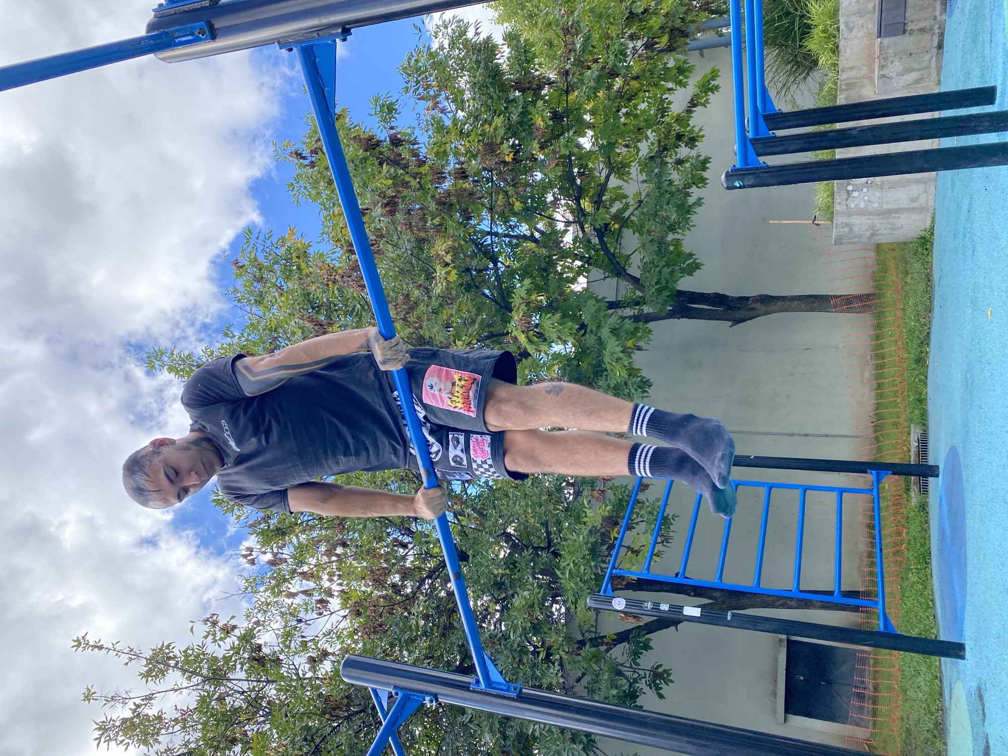 A man is exercising on a blue metal pull-up bar structure in what appears to be a park or outdoor gym. The man, likely in his late 20s to 40s, wears a dark-colored t-shirt, patterned shorts, and dark socks with light-colored stripes. He is gripping the pull-up bar with both hands, his body suspended in a horizontal position.
The structure is situated in a park-like environment with green grass and trees visible in the background. The sky is blue with scattered white clouds. A concrete wall is partially visible, and the ground appears to be a blue painted surface.
The location is likely Buenos Aires, Argentina.