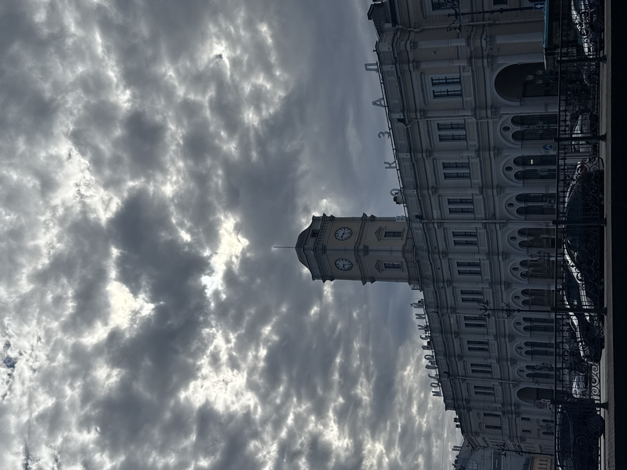The image displays a grand, classical-style building under a dramatic, heavily overcast sky, oriented as if viewed from across a street or elevated position. The setting is an urban environment, identified as Saint Petersburg, Russia, by the architectural style and visible text.
The primary subject is a large, multi-story building featuring a prominent clock tower centrally located on its roofline. The building has a light-colored facade, numerous arched windows, and ornate details characteristic of 19th-century European architecture. The clock tower itself is square, topped with a dome, and clearly displays at least two circular clock faces.
Horizontally arranged on the building's facade, to the left of the clock tower, is the Cyrillic text "МОСКОВСКИЙ" (Moskovsky). To the right of the clock tower, positioned slightly higher, the text "ВОКЗАЛ" (Vokzal) is visible, indicating the structure is the Moskovsky Vokzal (Moscow Railway Station).
The sky is filled with dense, textured dark gray clouds, with patches of brighter light breaking through, suggesting a cloudy daytime scene. The weather appears somber or majestic due to the dramatic cloud cover. In the immediate foreground, a section of an ornate black wrought-iron railing is visible, hinting at the photographer's vantage point. No people or specific activities are discernible in the static architectural scene.