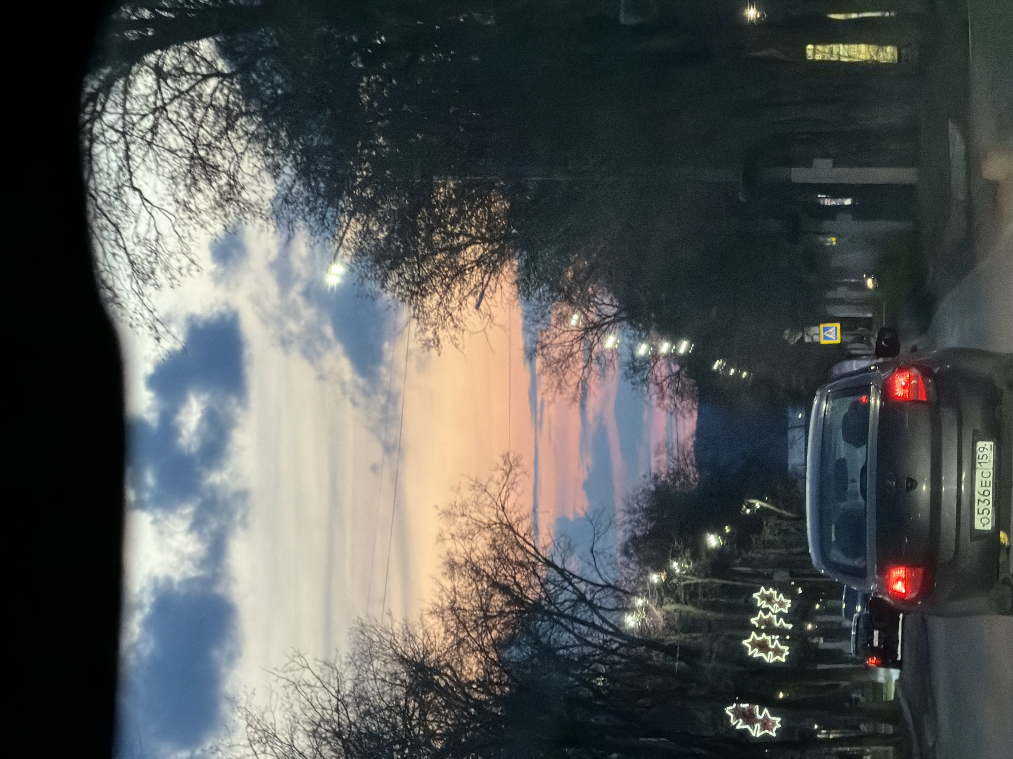 The image, best viewed horizontally despite its original vertical orientation, depicts a twilight street scene, likely captured from within a vehicle. The sky dominates the upper portion, showcasing a beautiful gradient from soft blue and white clouds on the left to warm orange and pink hues on the right, indicating either sunset or sunrise. Bare tree branches, silhouetted against the colorful backdrop, frame the scene, suggesting a late autumn or winter setting.
A road runs through the middle and right side of the image, illuminated by streetlights that cast bright glows. A grey car is prominently featured in the right foreground, viewed from behind, with its red taillights illuminated. Behind it, another vehicle is partially visible. Along the right side of the street, star-shaped light decorations are visible, suggesting a festive holiday season. Further down the road, a yellow pedestrian crossing sign can be discerned.
The license plate on the grey car reads "O 536 EC 159," with "159" being the region code for Perm Krai, Russia, confirming the location context of Chaikovsky. The combination of natural twilight, artificial street lighting, and festive decorations creates a peaceful yet subtly festive atmosphere, depicting a serene moment during an evening drive. The dark edges on the left suggest the view is framed by a car window or interior.