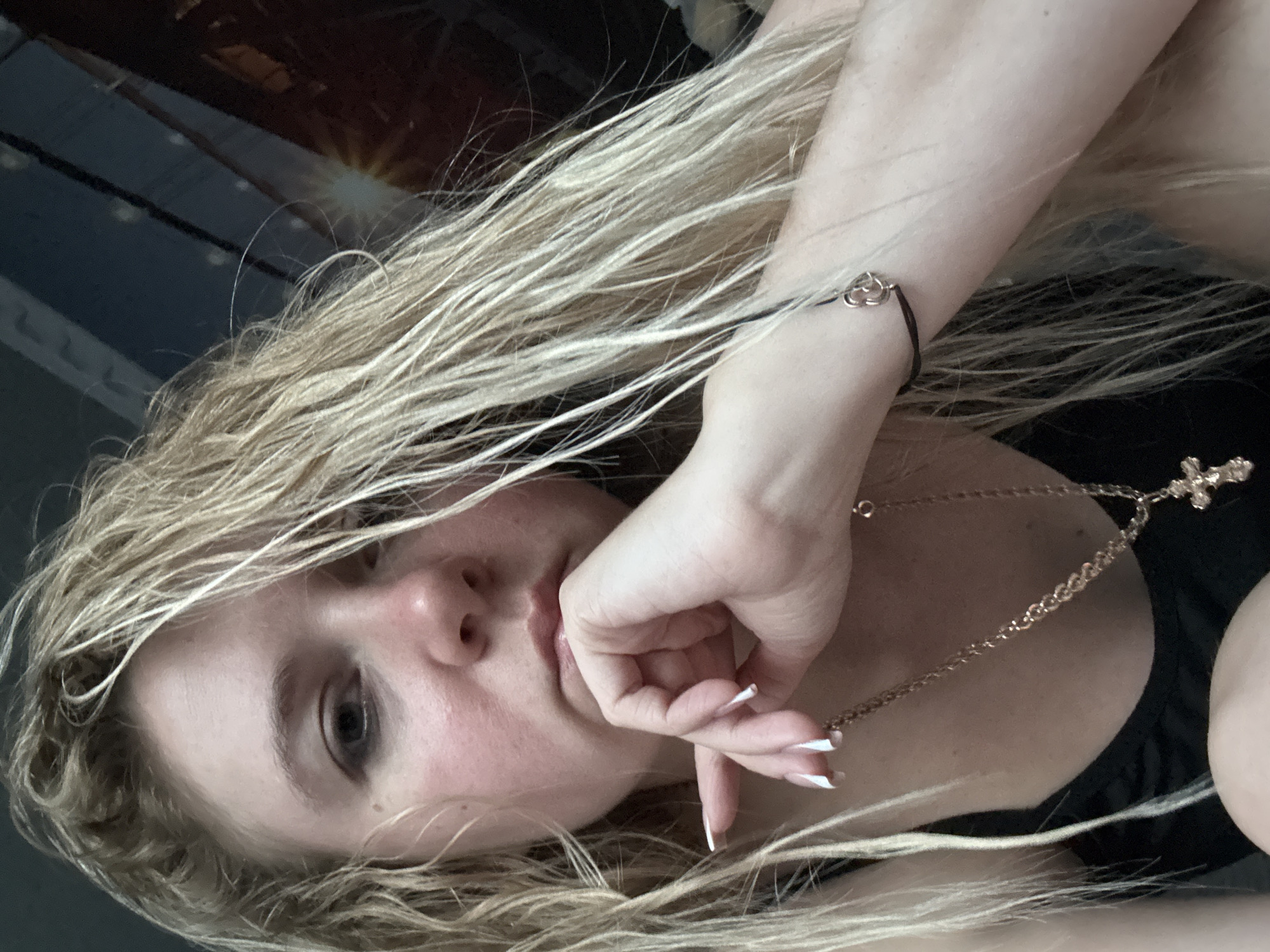A woman with blonde hair is captured in a close-up selfie. Her hand is resting on her chin, and she is looking at the camera with a contemplative expression. She is wearing a black tank top, a gold chain necklace with a cross pendant, and a black bracelet with a small, delicate charm. The background is dark and appears to be a wall with some indistinct patterns. The lighting is dim, creating shadows on her face.